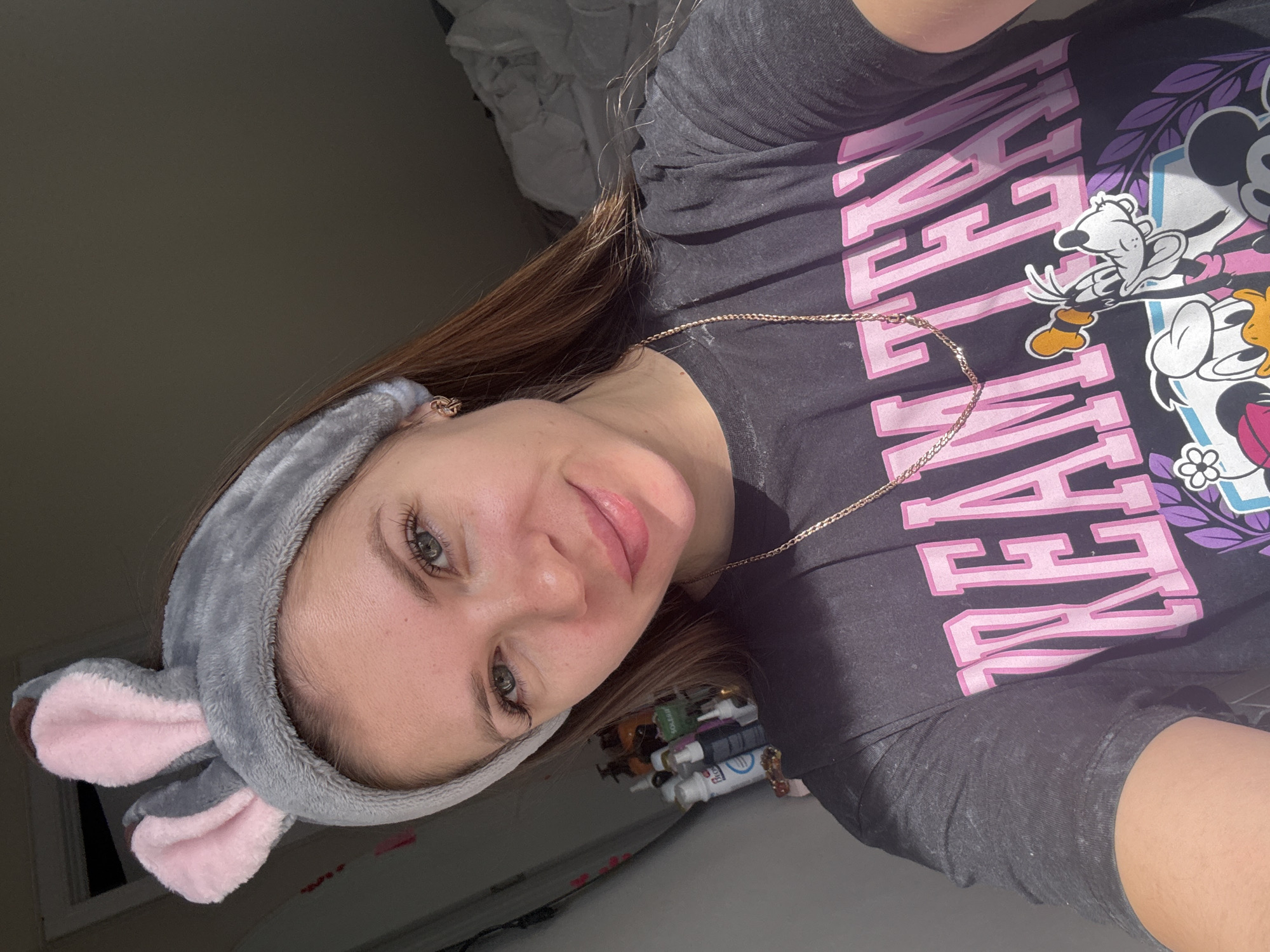 This image depicts a headshot-style selfie of a female individual indoors in Batumi, Georgia. The subject, positioned centrally, has light brown hair, light-colored eyes, and a fair complexion, showing a subtle smile. She is wearing a grey plush headband with pink-lined bunny ears. Her attire consists of a dark grey, short-sleeved t-shirt featuring pink block lettering that spells "TEAM" and a graphic below it showing animated characters including Goofy, Donald Duck, and Mickey Mouse. A thin, gold-colored chain necklace is visible around her neck, and she wears small, gold-colored earrings.
To the subject's left, a collection of bottles and containers, likely personal care products or toiletries, are visible on a surface, illuminated by a strong light source entering from the left side of the frame. To her right, a portion of crumpled, light-colored fabric, possibly bedding, is visible in shadow. The background suggests an indoor, domestic setting, potentially a bedroom or bathroom. The lighting creates a clear delineation between the well-lit left side and the shadowed right side of the image.