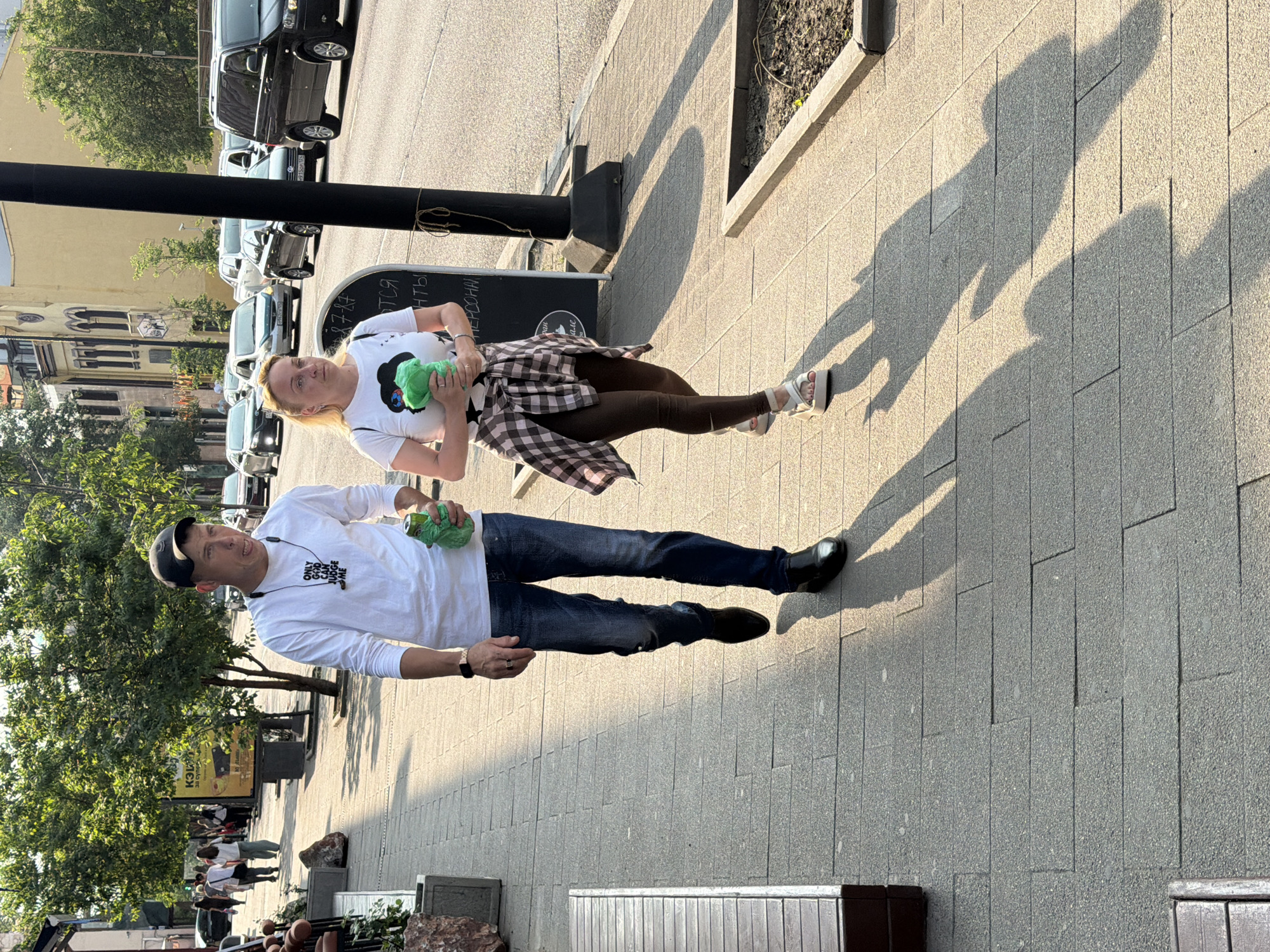 A man and a woman are walking on a sidewalk in Krasnoyarsk, Russia. The man is wearing a white long-sleeved shirt, blue jeans, and black shoes. He has a black baseball cap on and is holding a green object in his right hand. The woman is wearing a white t-shirt with a black graphic, brown leggings, and white sandals. She has a plaid shirt tied around her waist and is holding a green object in her left hand. They are walking towards the viewer. The sidewalk is paved with grey stones and is lined with trees. In the background, there are buildings and cars on a street. The sun is casting long shadows on the sidewalk.