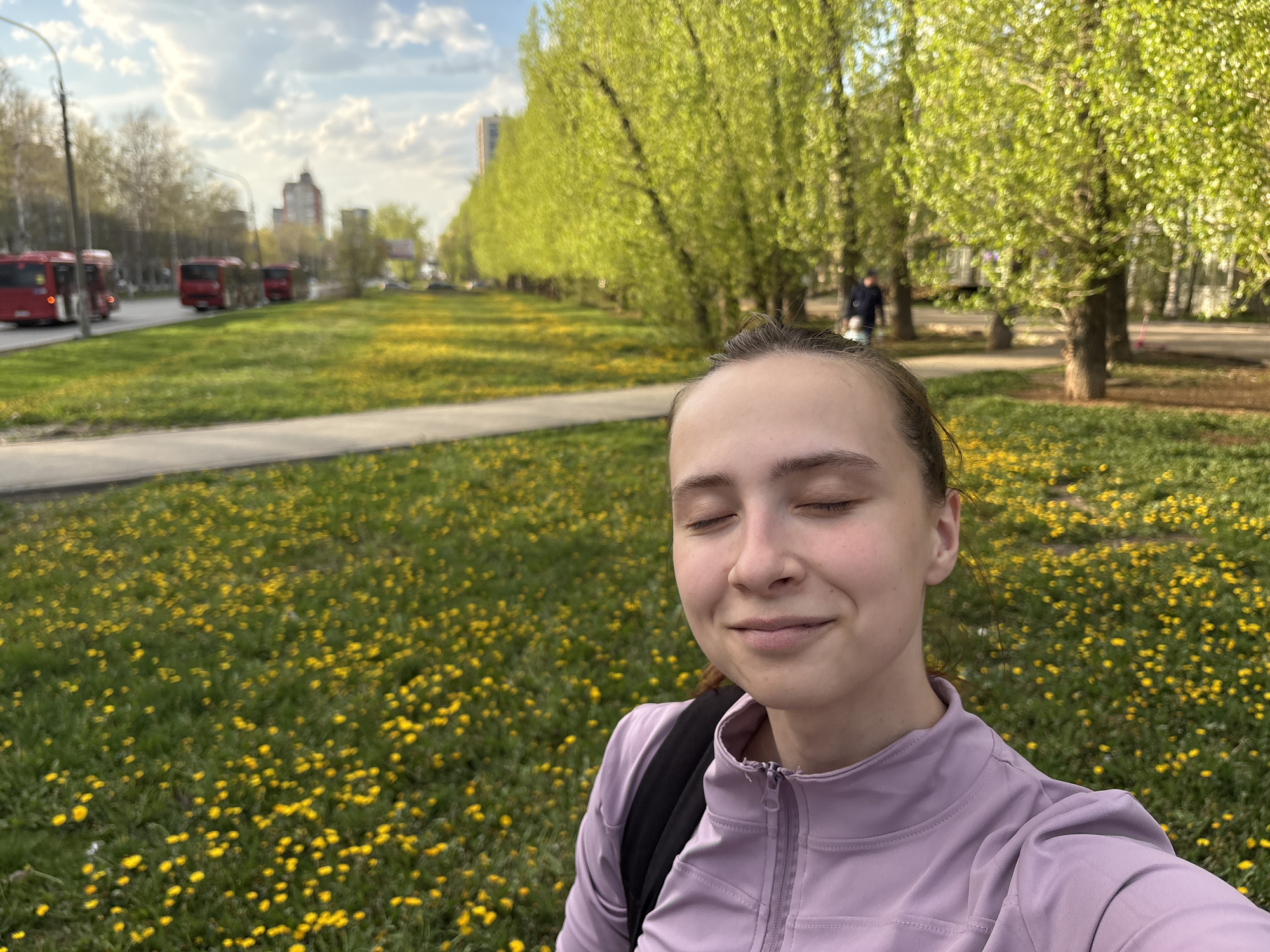 A young woman with fair skin, wearing a lavender zip-up jacket and a black backpack, is taking a selfie outdoors. Her eyes are closed, and she is smiling slightly. The background consists of a green grassy area filled with yellow dandelions. Behind the grassy area, a row of trees with light green foliage lines a walkway. Further back, there is a road with red buses, and buildings are visible in the distance under a partly cloudy sky. Based on the architecture, buses, and flora, the location is identified as being in the city of Perm, Russia.