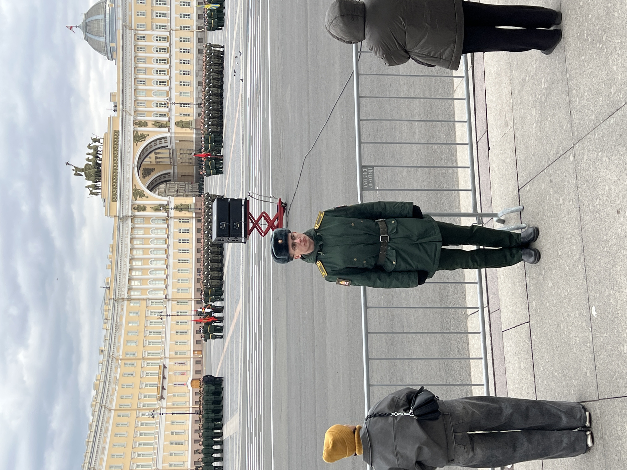 The image captures a military event or parade rehearsal in a large urban square, likely Palace Square in St. Petersburg, Russia. In the immediate foreground, a uniformed male soldier in a dark green overcoat and a black fur hat stands behind a metal crowd control barrier, looking directly at the camera with a neutral expression. To his right and left, partially visible from behind, are two civilian onlookers. One wears a dark hooded jacket and black pants, while the other has a dark jacket, dark pants, and a yellow beanie, with a black shoulder bag.
Behind the barrier and the central soldier, a wide, paved parade ground extends into the distance, marked with white lines. Numerous soldiers in dark green uniforms are visible in formation, standing in neat rows. Some soldiers closer to the historical buildings carry red flags or banners. To the left of the foreground soldier, a set of speakers on a red stand is positioned on the edge of the parade ground.
In the background, magnificent neoclassical architecture dominates the scene. A grand yellow and white building with numerous windows and columns stretches across the frame, featuring a prominent archway topped with a large bronze sculpture of a chariot pulled by horses (part of the General Staff Building). Further to the left, another large building section with a dome is visible. The sky is overcast, suggesting a cool, bright daytime. On the metal barrier to the right, partially visible Russian text reads "ВХОД ДЛЯ УЧАСТНИКОВ" (ENTRANCE FOR PARTICIPANTS).