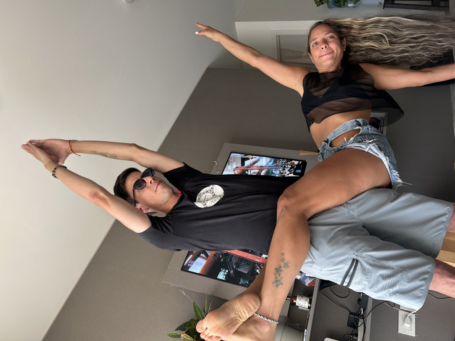 The image captures two people, a man and a woman, striking a playful pose in what appears to be a modern indoor setting, possibly a living room or bedroom. The photograph is taken from a low angle and is rotated approximately 90 degrees clockwise, making the light-colored ceiling appear as a vertical wall on the left and the actual floor as a vertical wall on the right.
The man is positioned horizontally across the frame, seemingly lying on his back or reclining, with his arms stretched out above his head, palms together, mimicking a flying or superhero stance. He is wearing dark sunglasses, a black short-sleeved t-shirt with a white circular graphic print on the front, and light grey athletic shorts. On his left wrist, he wears a red string bracelet and a dark beaded bracelet. Tattoos are visible on both of his forearms. His expression, partly obscured by the sunglasses, appears relaxed.
The woman is perched on the man's torso and lap, also in a horizontal orientation, mirroring his outstretched arms, as if flying or balancing. She is looking directly at the camera with a slight smile or smirk. Her long, wavy blonde hair flows out behind her to the right. She is wearing a sheer black cropped top over a black bra and distressed light-wash denim shorts with a cutout detail on the left thigh. A green, vine-like tattoo is visible on her left calf/ankle, and she wears a silver-colored anklet on her left ankle. Her bare feet are visible, with the sole of her left foot prominent in the foreground.
In the background, behind the man's head and the woman's legs, a large flat-screen television or monitor is visible, displaying an image that appears to be from a video game or an action scene, with warm orange and dark tones. Below the screen, a small potted plant with variegated leaves is present. The overall lighting suggests it is daytime, with ambient light illuminating the room. The mood conveyed is lighthearted and fun, typical of candid or posed personal photos. No discernible text is visible in the image.
