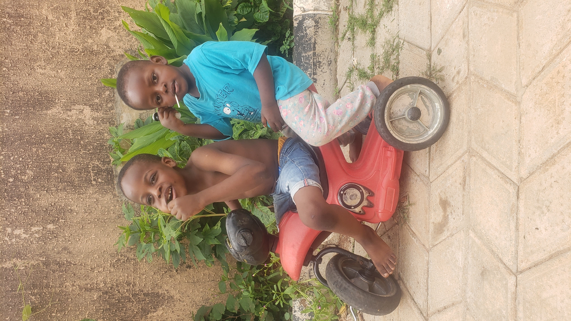 Two young boys are sitting on a red toy motorcycle in what appears to be a backyard or outdoor play area. The boy in front, with his chest bare, is wearing denim shorts and has his hand to his mouth. The boy behind him, wearing a blue t-shirt with graphics and patterned leggings, is holding a lollipop. They are positioned on a paved surface, with a textured wall and lush green foliage in the background. The scene is brightly lit, suggesting daytime. The location is identified as Sabon Tasha, Nigeria.