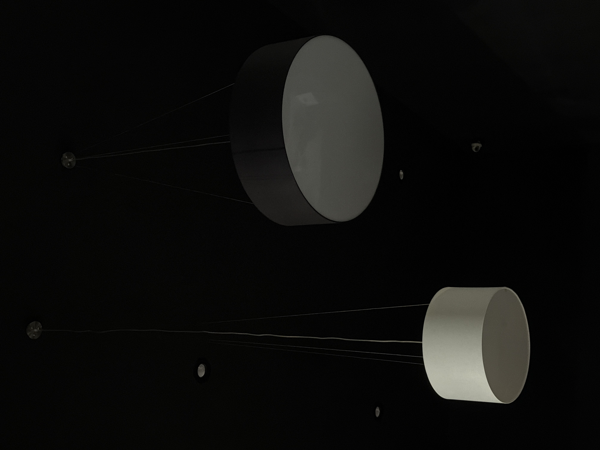 The image shows two cylindrical light fixtures suspended from the ceiling by cables in a dark interior space. The top fixture is larger, with a dark outer casing and a light gray face. The lower fixture is smaller and entirely light gray. Small circular objects are visible on the ceiling, suggesting mounting points or fixtures. The environment is generally dark, with the primary light source being the fixtures themselves. There is no indication of the location being Izhevsk, Russia, or any people present.