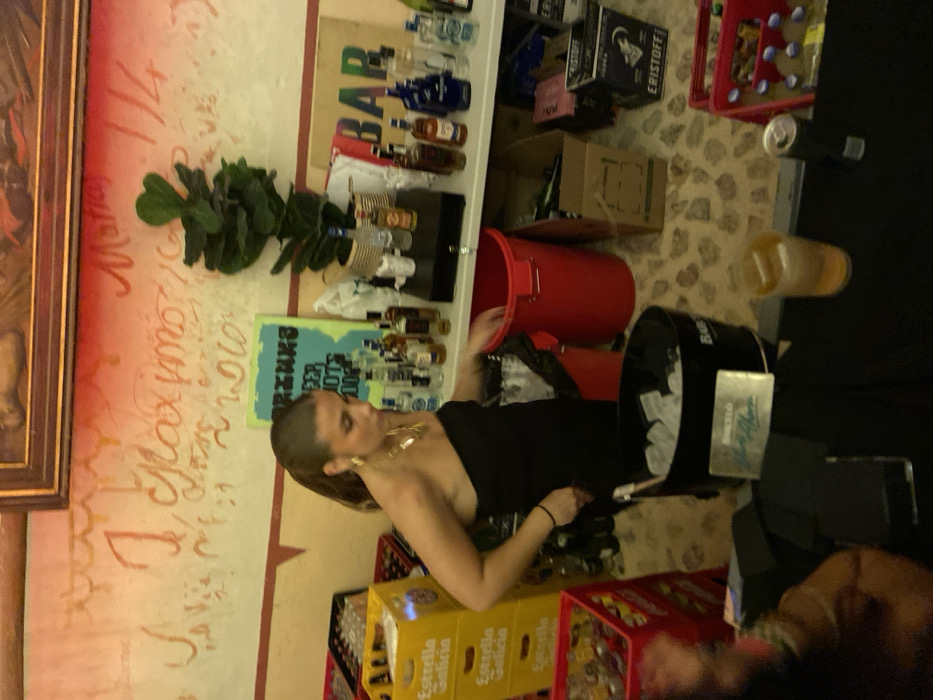 The scene is a bar or restaurant in Cabanillas, Spain, at the coordinates 40.977 latitude, -4.032 longitude. A woman, wearing a black strapless dress and gold jewelry, stands beside a bar counter with an open red trash bin. She is looking into the bin. On the counter, there is a bucket with ice, a glass of liquid, a can of drink, and several boxes of alcoholic beverages. Behind the counter is a shelf stocked with bottles of liquor. The wall above the counter features some handwritten writing and an advertisement for "Premios." The floor is a mosaic of beige stone tiles.