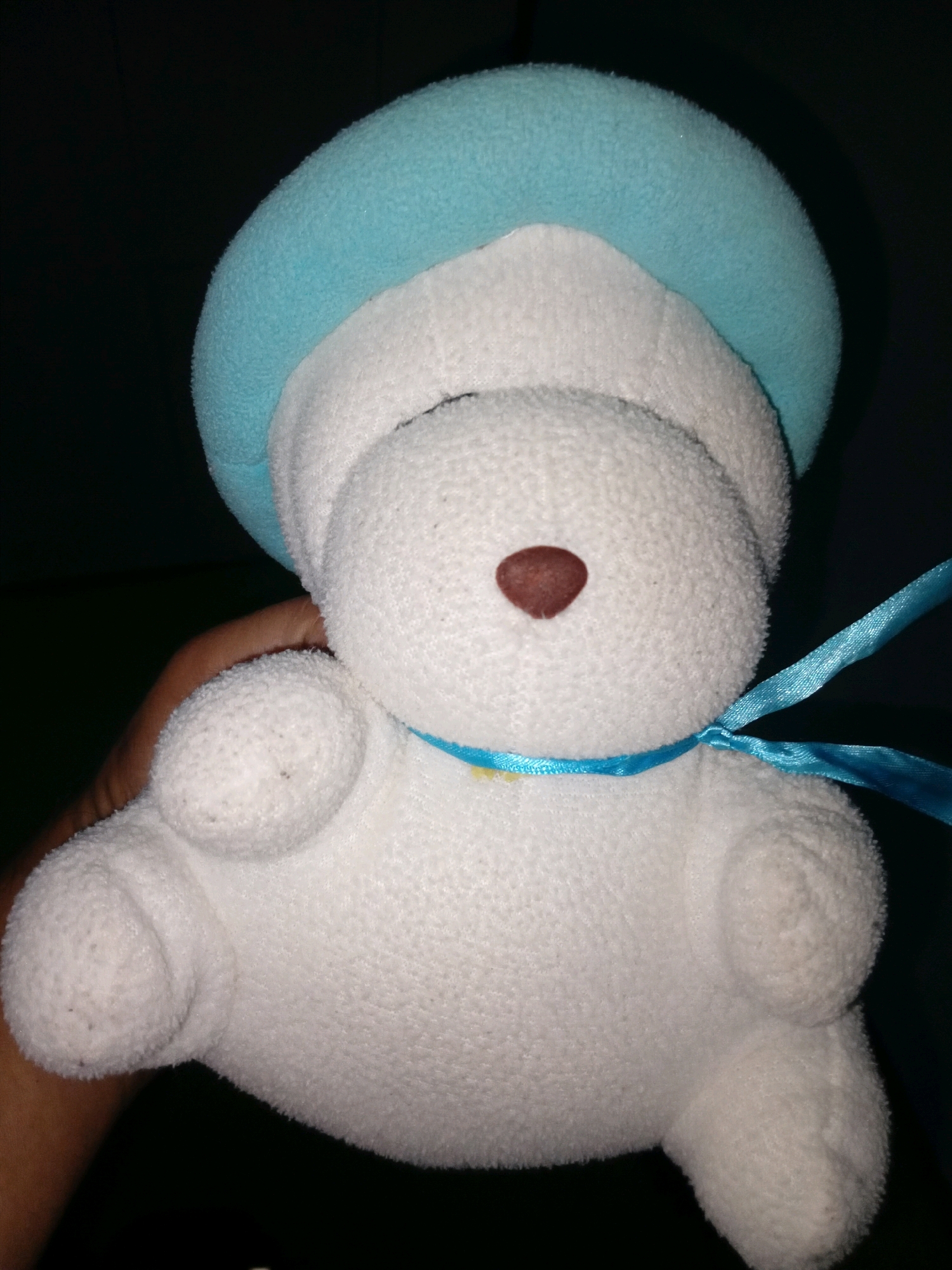 A close-up, full-body shot features a white, plush teddy bear wearing a light blue beanie. The bear is being held by a hand, parts of which are visible on the left side of the frame. A thin blue ribbon is tied around the bear's neck. The background is dark and out of focus. The scene is indoors, possibly in a dimly lit room in Gebang, Indonesia.