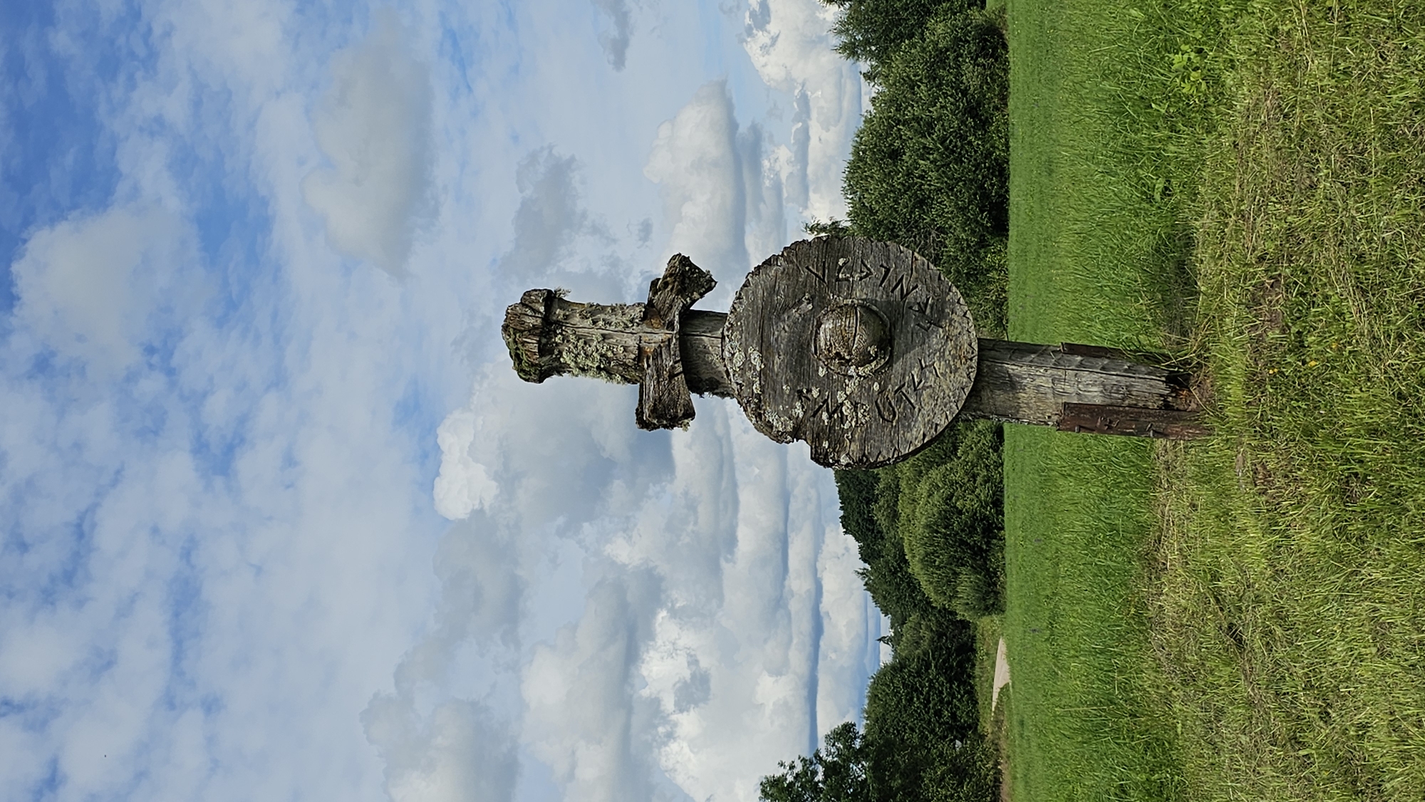 A weathered, wooden object, resembling a sword or a large cross with a circular shield, stands prominently in a grassy field. The object is made of rough-hewn wood, showing signs of age and exposure to the elements, with moss and lichen growing on its surface. The circular part, positioned below the cross-like top, is carved with symbols or runes and has a central, circular indentation. The object is mounted on a thicker wooden post, which is reinforced at its base with metal plates. Behind the wooden object, a green field stretches out, leading to a treeline of dense, lush foliage. The sky above is a vibrant blue, dotted with fluffy white clouds. The image appears to be taken outdoors, in a rural or natural setting. This object is located in Embūte, Latvia, and is a significant cultural and historical landmark, often referred to as the Embūte Stone or the Embūte Wooden Idol.