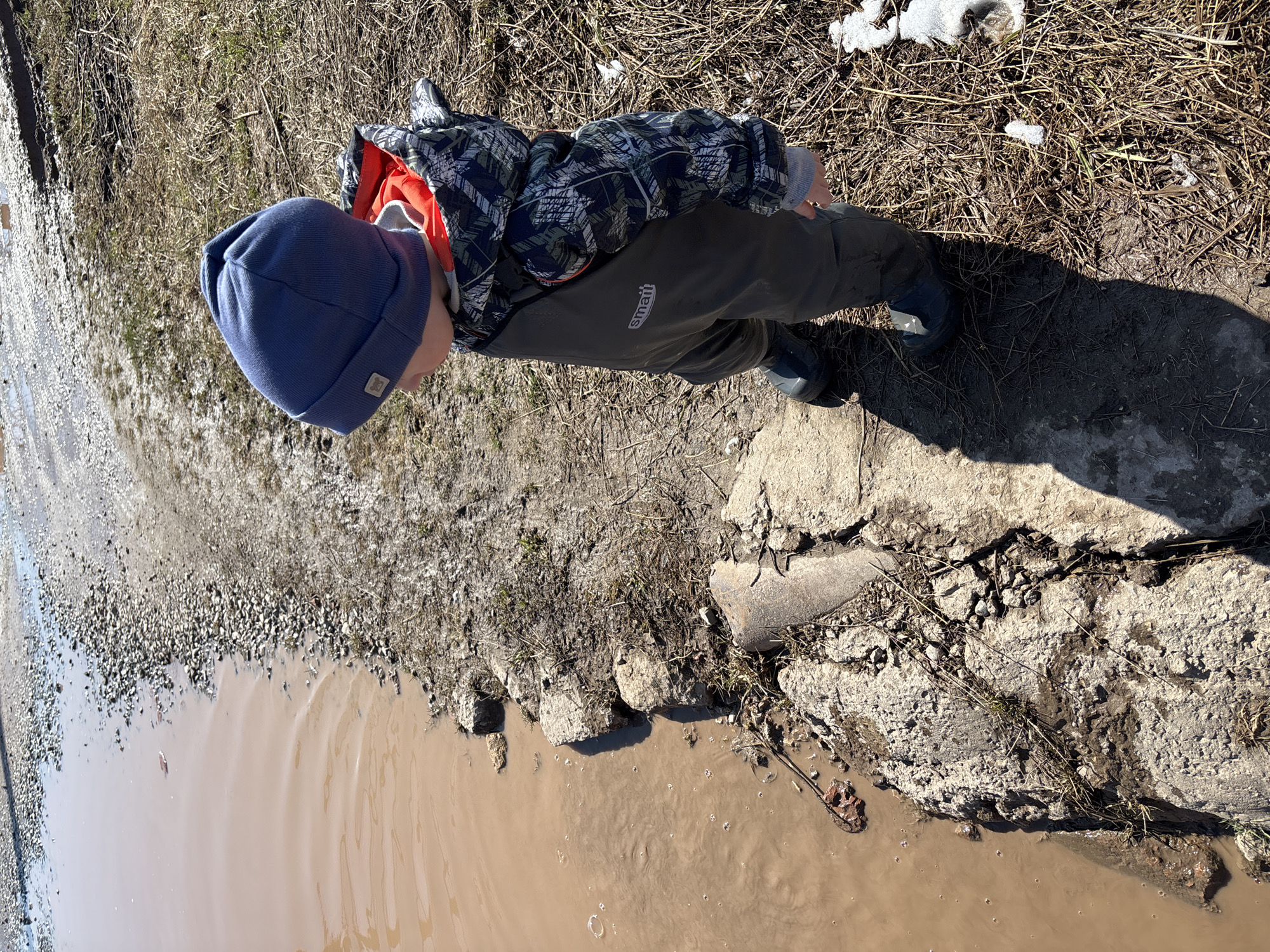 A young child is standing near a body of muddy water at an unspecified location in Ferma, Russia. The child is wearing a blue knit hat, a patterned jacket with an orange lining, and dark green overalls, along with dark rubber boots. The ground around the water is a mix of gravel, grass, bare earth, and broken concrete. There is a small patch of snow in the background. The muddy water has bubbles on the surface. A concrete pipe sits partially submerged in the water.