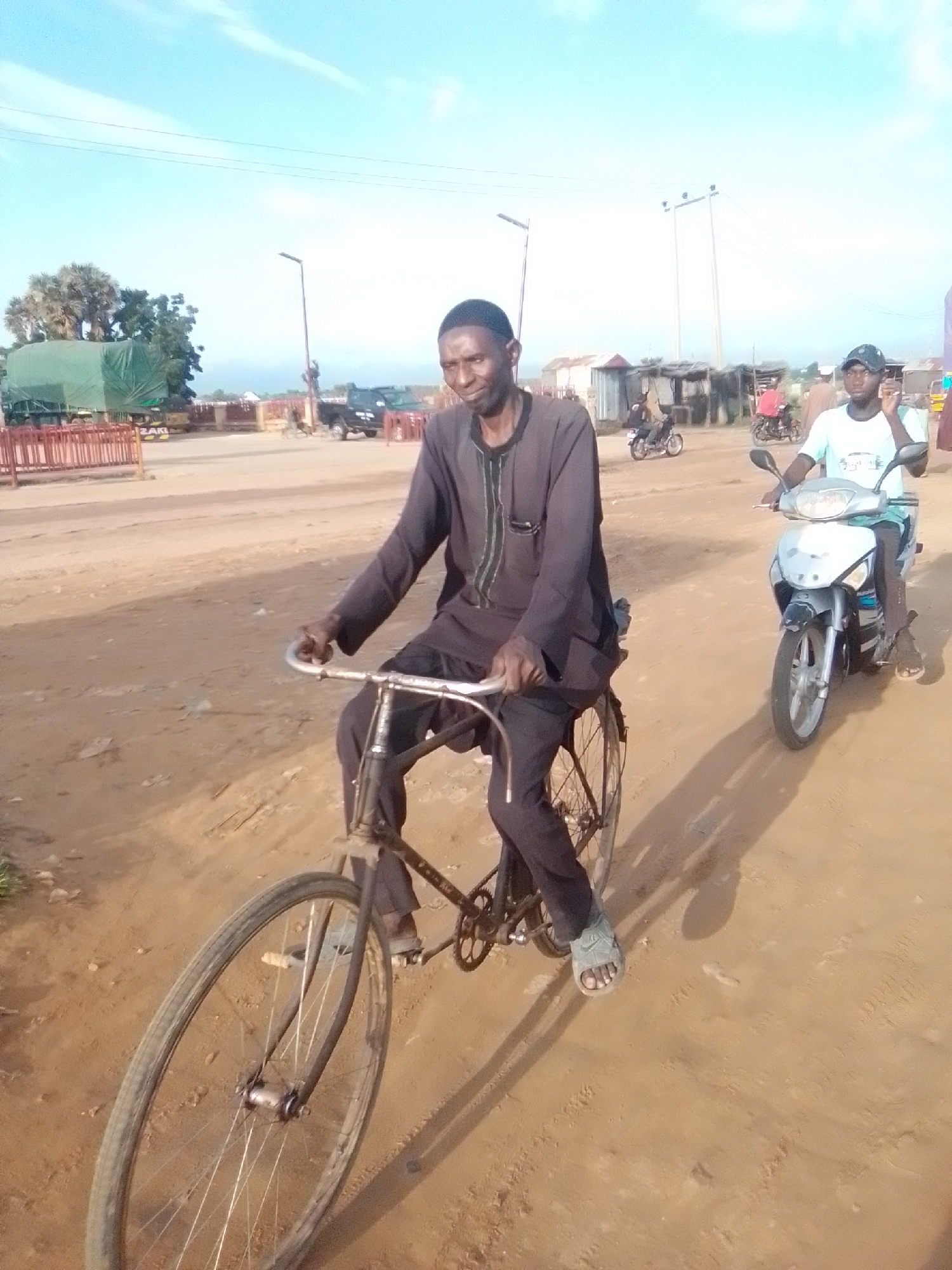 The image captures a street scene in Kwadon, Nigeria. In the foreground, a man is riding a bicycle, wearing a dark traditional outfit. His left foot, clad in a sandal, is visible on the pedal. Behind him, another person is riding a scooter, wearing a light-colored t-shirt and a cap. The environment is dusty and unpaved, with a few structures and trees visible in the background. The sky is clear and blue with some faint clouds. There are other vehicles and people in the distance, suggesting a bustling area.
