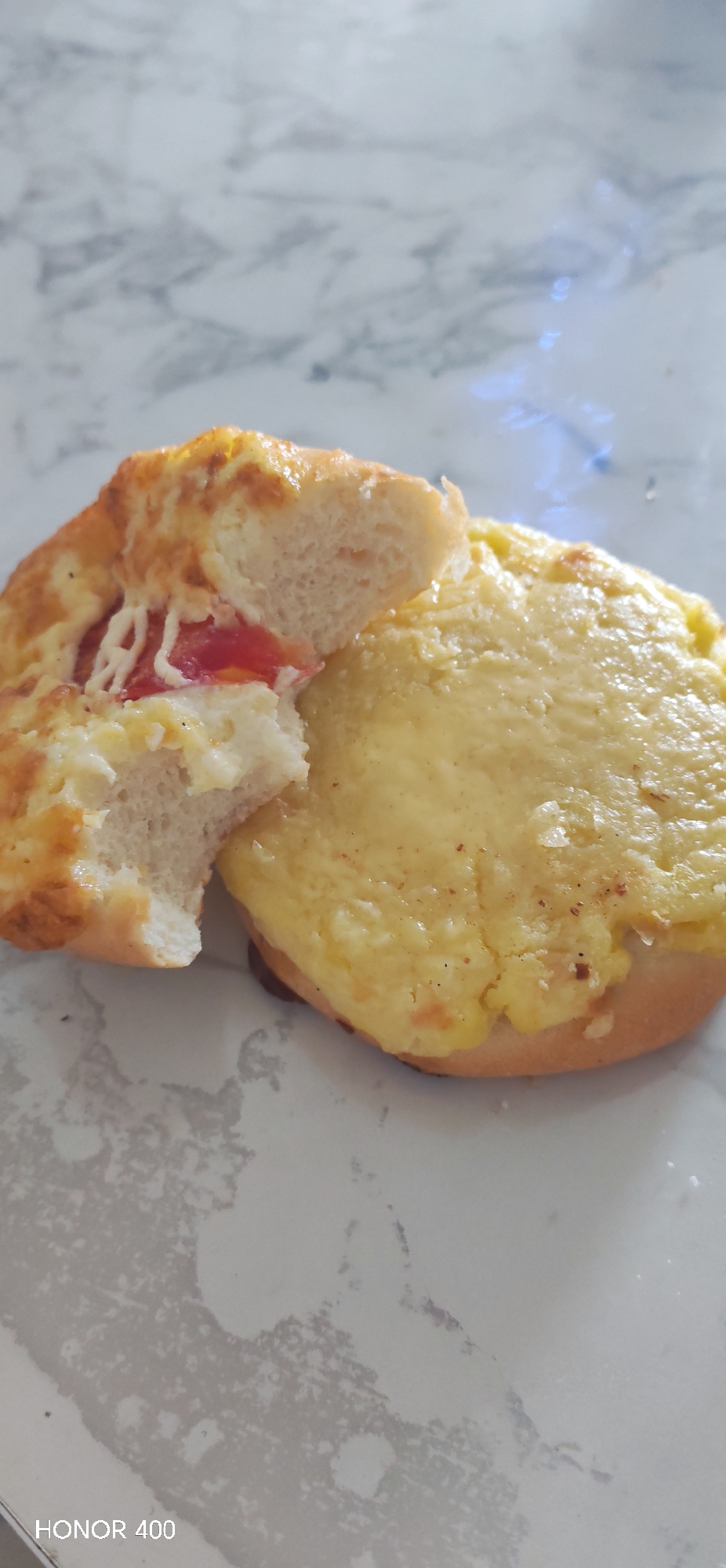 On a marble surface, two pastries are displayed. One is cut in half, revealing a bread-like interior with a layer of tomato and cheese. The other pastry is covered in a thick layer of melted cheese with visible browned spots. The location is not discernible from the image.