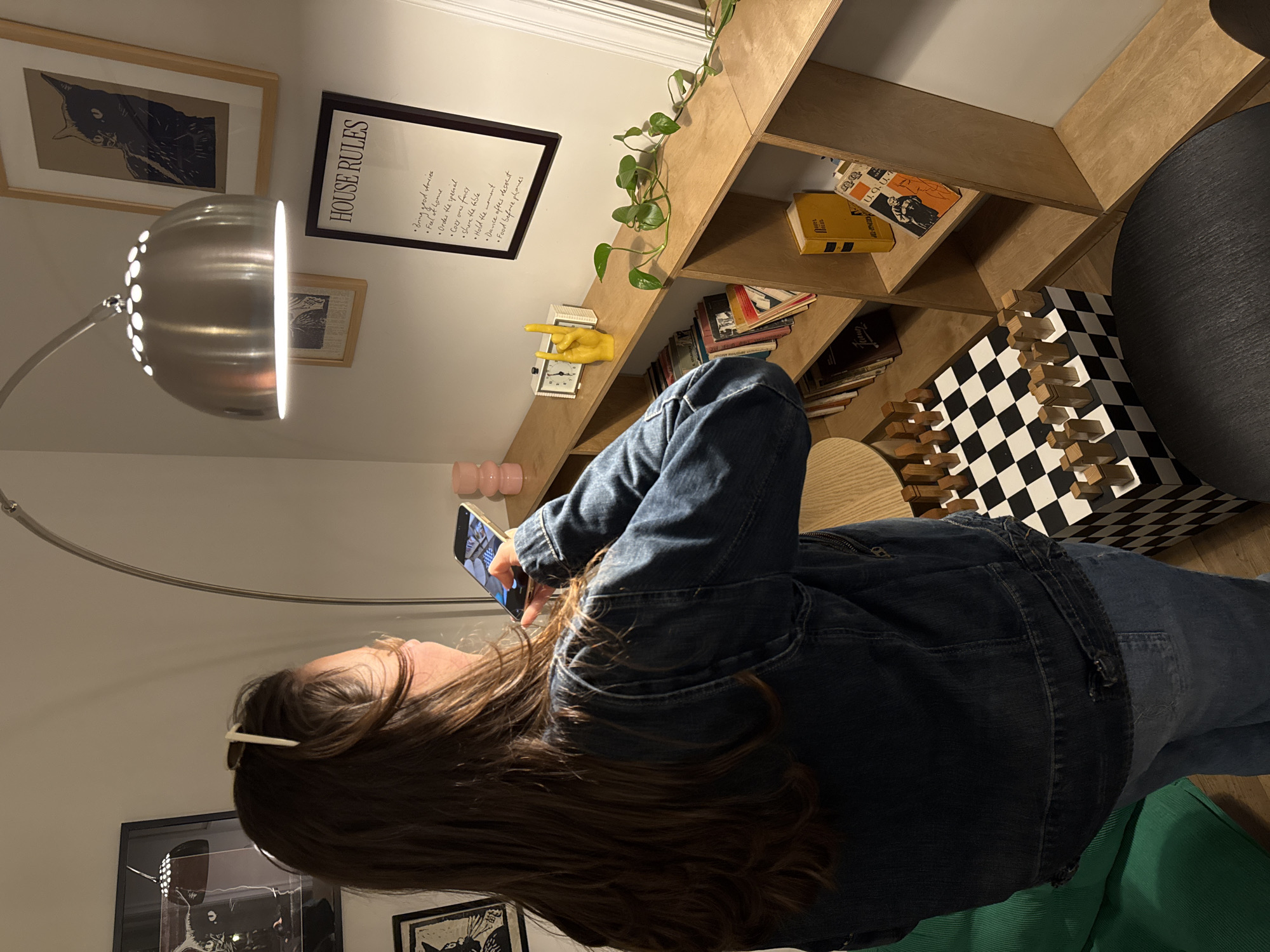 A woman with long brown hair, wearing a denim jacket and jeans, is standing and holding a smartphone, appearing to take a picture or use the phone in a room with a homey and artistic decor. The room features a light wood bookshelf filled with books, a yellow clock, and greenery. There is a chessboard with wooden pieces, and framed artwork adorns the walls, including a "House Rules" poster. A modern arched floor lamp provides lighting. The location is not discernible from the image.