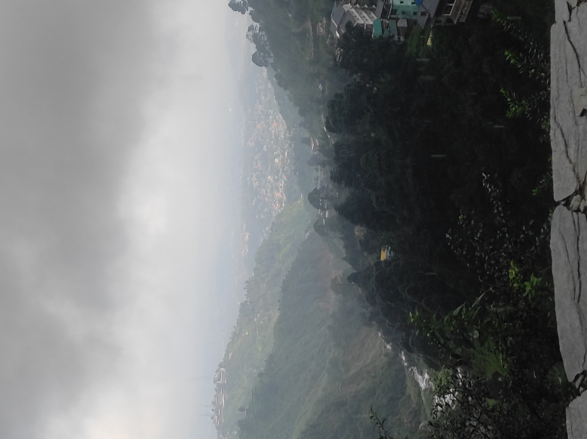 The image depicts a panoramic view of a valley town, likely Bhagsu, India, shrouded in a thick layer of mist and clouds. The landscape is characterized by steep, verdant hillsides dotted with buildings and dense tree cover. In the foreground, a portion of a stone wall or pathway is visible, suggesting an elevated viewpoint. The middle ground features a winding river or stream flowing through the valley, flanked by dark green trees. Further into the valley, a sprawling town with numerous buildings is discernible, its details softened by the atmospheric conditions. Several multi-story buildings, some with visible balconies and roofs of varying colors, are scattered along the hillsides, particularly towards the right side of the image. The overall impression is one of a serene, overcast day in a mountainous region, with a significant urban settlement nestled within the natural landscape.