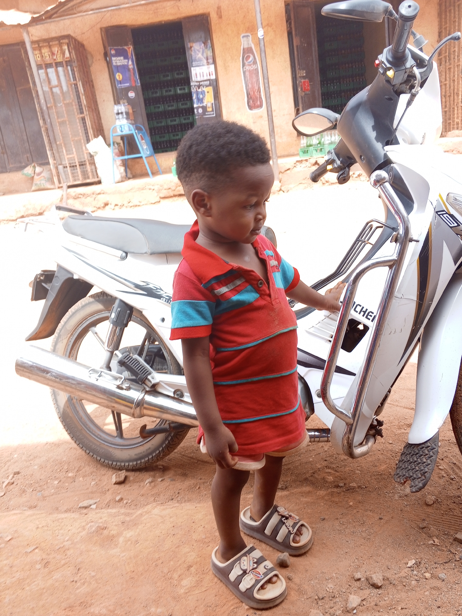 A young child stands beside a white motorcycle on a dirt surface in Yelwa, Nigeria. The child wears a red and blue striped shirt, red shorts, and sandals. The motorcycle is parked in front of a building with open doors revealing shelves stocked with beverages. Posters and signage are visible on the building's exterior.