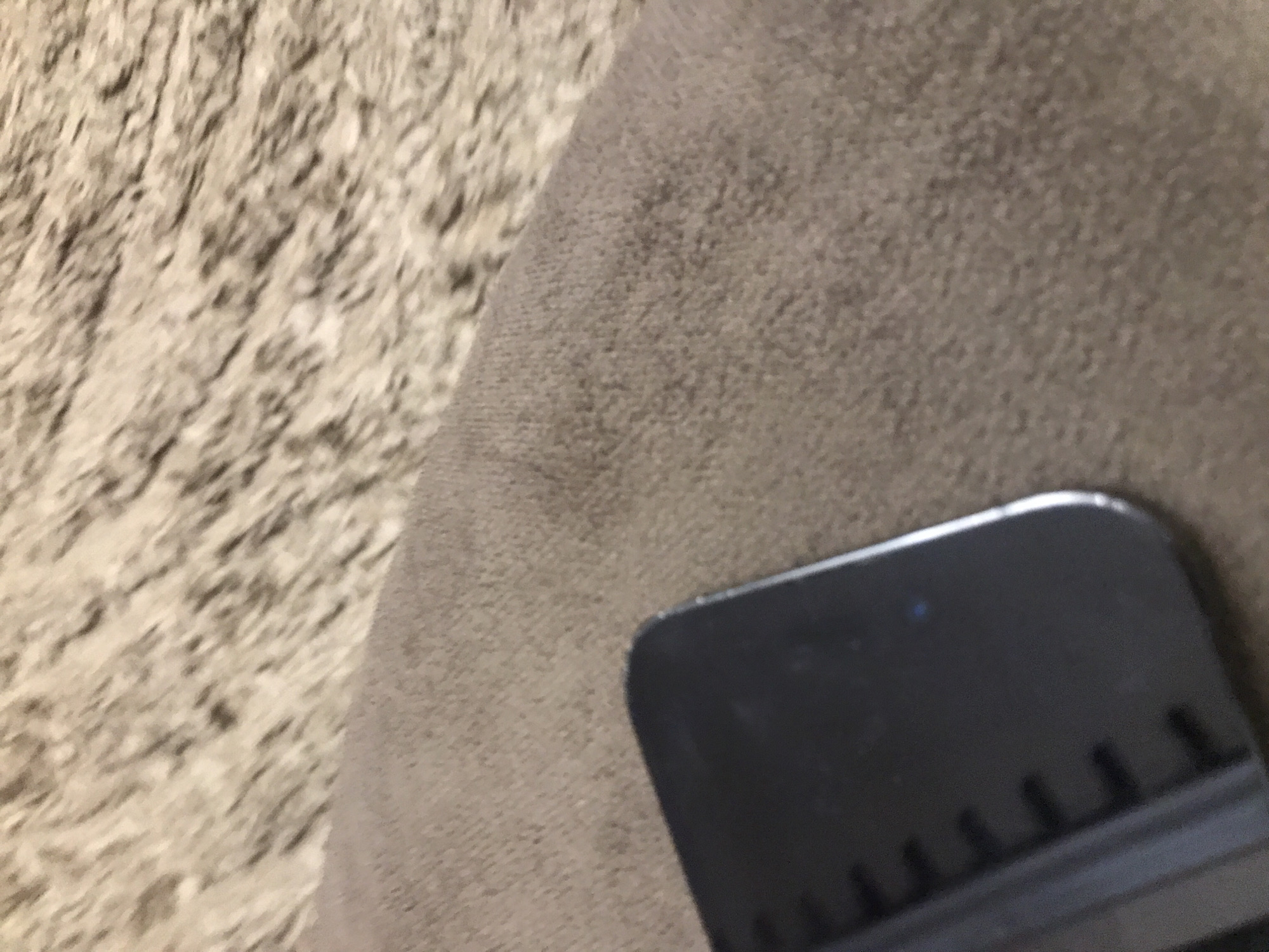 The image shows a close-up of a surface with a black object partially visible in the lower right corner. The surface to the left is a textured, light brown material, possibly carpet or a similar fabric. The surface to the upper right is a smooth, light brown material. The black object appears to be a part of a smartphone. There is no information available to determine the location in Yerevan, Armenia or any details about people, subjects, actions, or interactions.