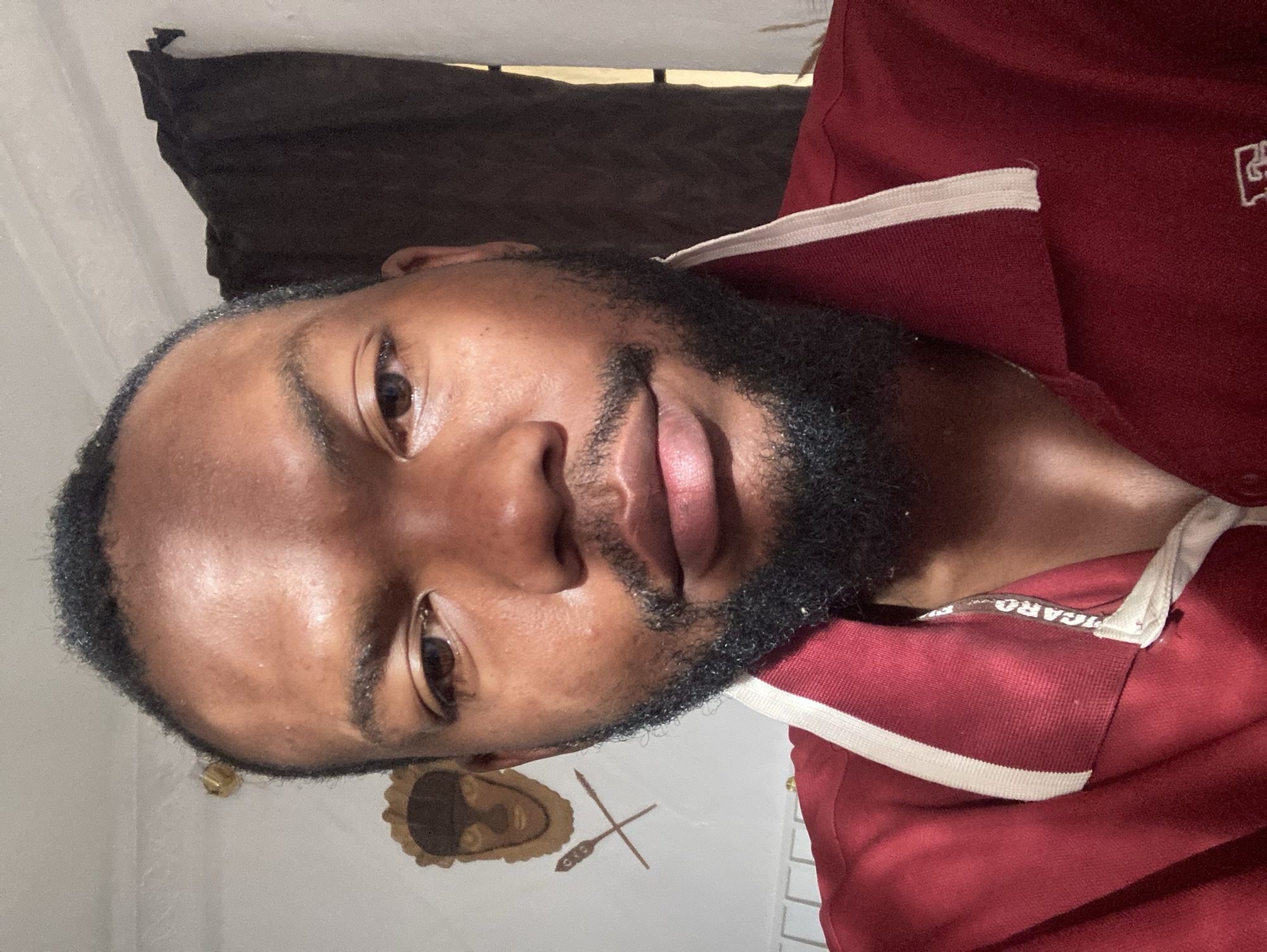 A portrait of a man with dark skin and a beard, wearing a maroon polo shirt with a white collar, is presented. The man is positioned at an angle, with his head tilted slightly. Behind him is a dark curtain, and the wall has artwork depicting a face and crossed spears. The collar of the polo shirt is visible and includes the printed word "PICARO." No information is present to establish the image's location in Jos, Nigeria.