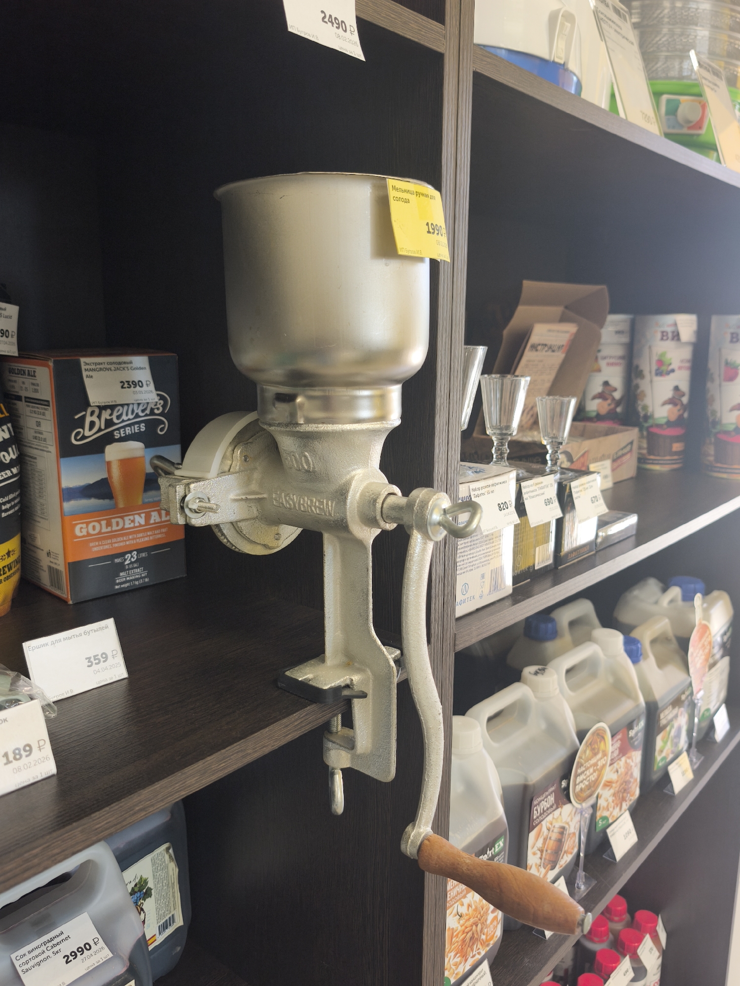 The image shows a hand-cranked grain mill mounted on a shelf in a store. The mill is made of metal and has a wooden handle. It is positioned on the second shelf from the top, among various products. To the left of the mill is a box of "Brewer's Series Golden Ale" extract. To the right of the mill, on the same shelf, are small glasses, boxes with prices, and cans of what appear to be beverage ingredients. The shelf below contains several large plastic containers, likely for liquids, and a bottom shelf with bottles. A price tag for the mill indicates 1990 rubles. Another price tag on the shelf below the mill is for a "Bottle brush" at 359 rubles. A larger price tag for a "Sauvignon Blanc grape extract" is visible on the bottom shelf at 2990 rubles. The overall environment appears to be a retail space, possibly a home brewing supply store. The lighting suggests an indoor setting with artificial lights. The image appears to be taken at a slight angle, looking up towards the shelves. The presence of Russian currency (rubles) and Cyrillic text on the product labels indicates the location is likely in Russia. Based on publicly available information, a store selling such items in Yoshkar-Ola, Russia, would be a plausible setting.