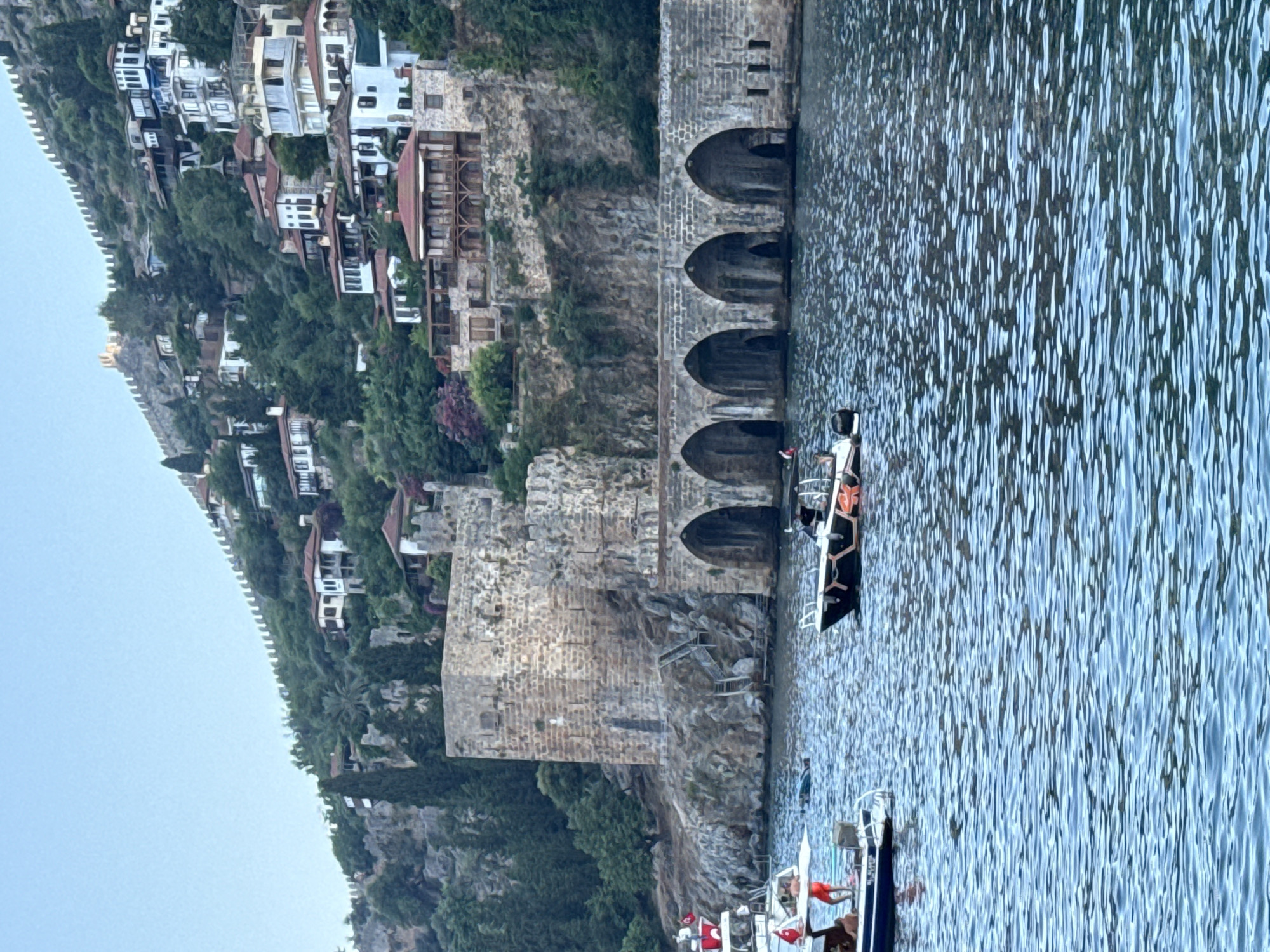 The image depicts a waterfront scene in Alanya, Turkey. The foreground is dominated by the blue, rippling water of the sea. A small boat with people on it, some wearing life vests, is visible in the water.
Behind the water, a significant stone structure with arched openings rises from the rocky shore. This appears to be part of a historic fortress or harbor wall. Further up the steep hillside, a cluster of buildings with white walls and red-tiled roofs are nestled amongst lush green trees. A stone wall runs along the crest of the hill, overlooking the sea and the buildings.
The scene suggests a daytime setting, possibly late afternoon or early evening, given the light and the calm water. The weather appears to be pleasant. The architecture and the natural landscape are characteristic of coastal Mediterranean regions.
There is no visible text in the image. The overall impression is one of a scenic and historic location, likely a popular tourist destination.
