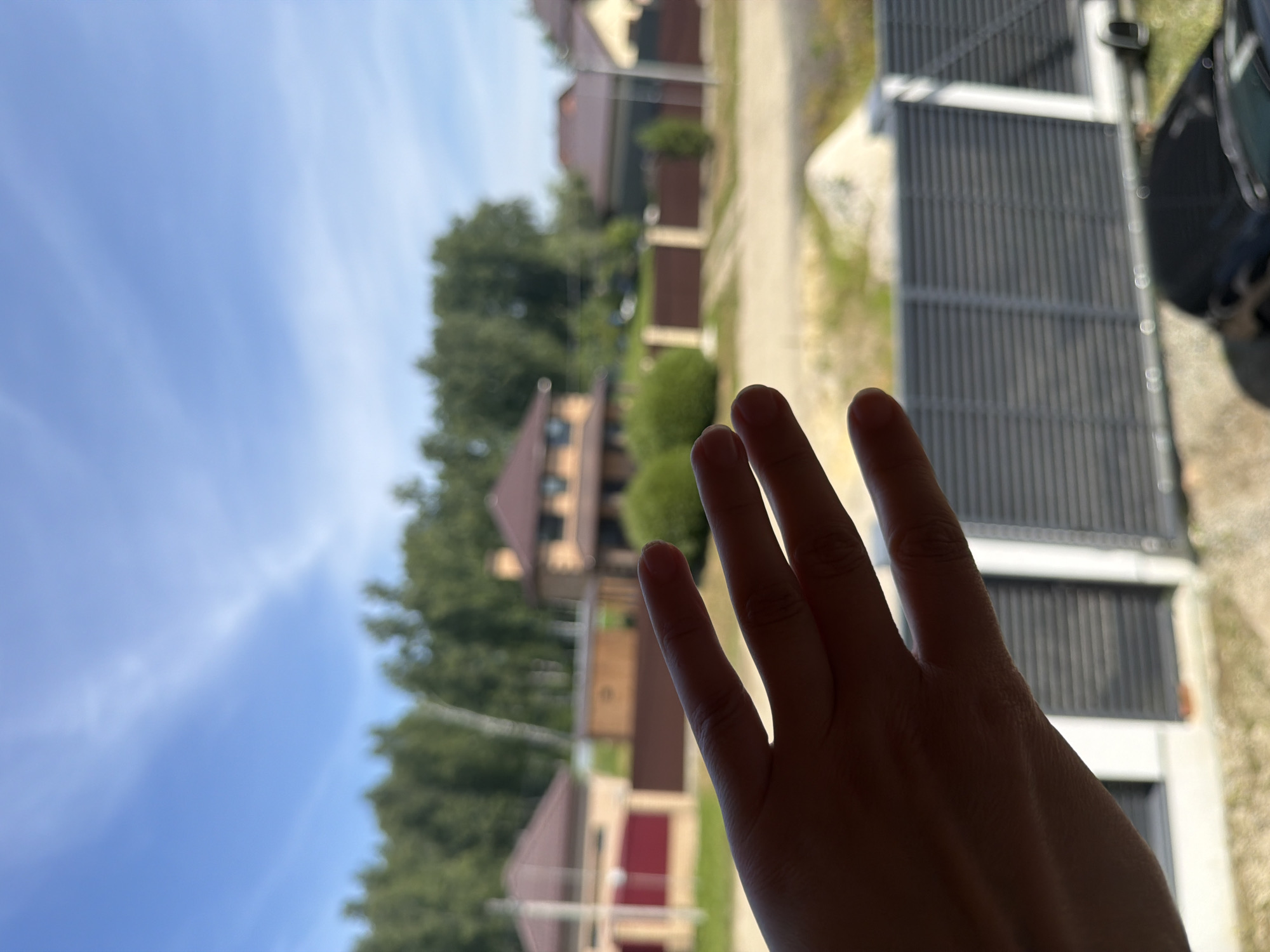 The image shows a close-up of a hand with fingers spread, partially obscuring a view of a suburban street scene. The hand is in the foreground and is dark in silhouette against a bright background. The background displays several houses with reddish-brown roofs, green trees, and a blue sky with wispy clouds. A dark-colored car is visible in the lower right of the frame. A gray, slatted fence runs horizontally across the middle ground. The overall setting appears to be a residential area in Malinovka, Russia, under a clear sky.