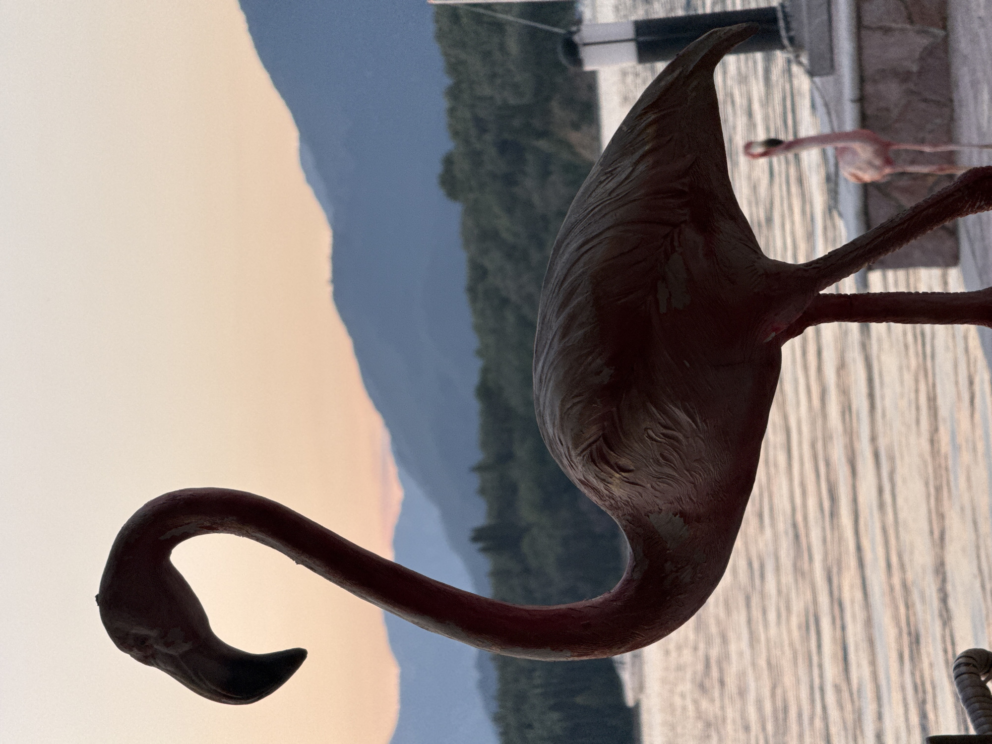 The image is a landscape with a pink flamingo statue in the foreground. The scene is likely near Bogišići, Montenegro, given the landscape context. Behind the flamingo is a body of water that appears to be a lake or bay, and beyond that are mountains covered with trees. The sky is a gradient of orange and pale blue, suggesting either sunset or sunrise. Another flamingo is visible in the background. The photo is taken from a low angle.