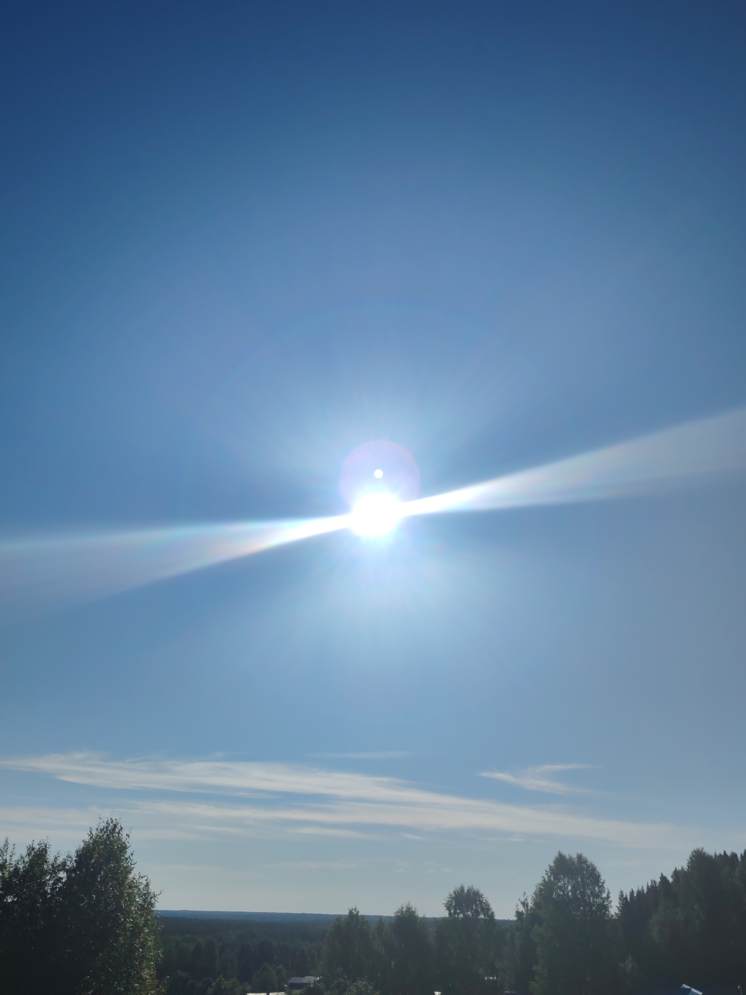 The image depicts a bright, clear day with a nearly cloudless blue sky dominating the upper portion. The sun is positioned centrally, emitting a strong glare and lens flare that extends horizontally across the frame, creating visible light rays. In the lower third of the image, a dense, dark green forest stretches across the horizon. The foreground features the tops of trees, some of which are lighter green and appear to be deciduous, while others are dark and coniferous. Distant structures, possibly houses or buildings, are faintly visible within the treeline. The overall scene suggests an outdoor, natural environment with a strong emphasis on the sunlight and sky. The location is identified as Чухлэм, Russia.