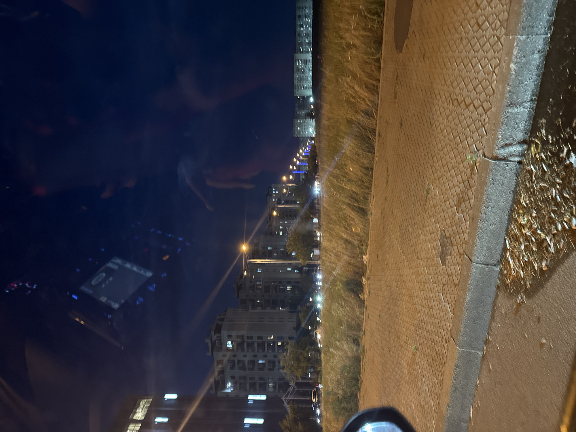 The image is a nighttime shot taken from inside a vehicle, looking out at a street scene. The sky is a deep blue. In the foreground, there is a paved walkway with interlocking bricks and a concrete curb. Dry grass and some fallen leaves are visible along the edge of the walkway. Beyond the walkway, there is a row of streetlights with yellow and blue lights. In the middle ground, several multi-story apartment buildings are illuminated, with many windows lit up. A line of cars, some with headlights on, are moving along the street. In the distance, more apartment buildings are visible. The scene appears to be in a residential area of a city. There are reflections on the glass of the vehicle, showing parts of the car's interior, including a dashboard screen and red lights.