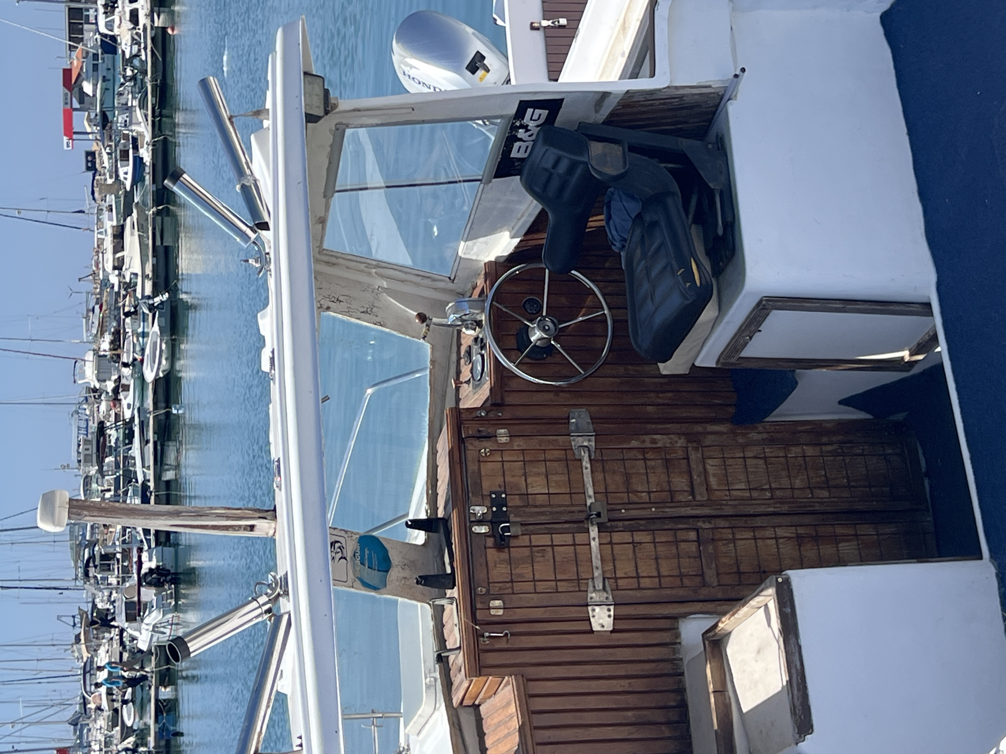 The image shows the interior of a boat's cockpit, with a steering wheel, seat, and a wooden cabin door. A Honda engine is visible in the background. Outside the boat, a marina filled with other boats can be seen, and beyond that, the city of Bar, Montenegro.