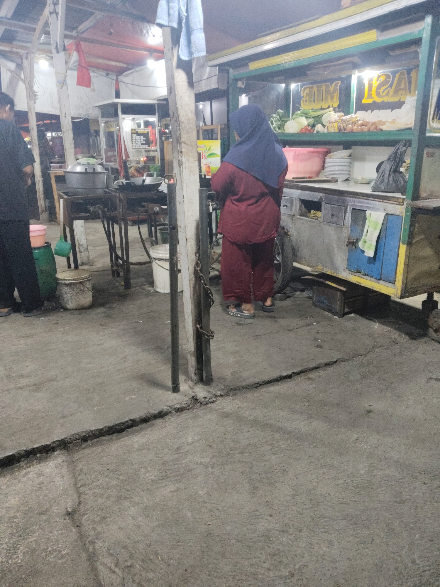The image depicts a street food stall in Subang, Indonesia. A woman wearing a blue hijab and a maroon outfit stands facing away from the camera, looking towards the stall. The stall itself is colorful, with yellow and blue accents, and displays various ingredients, including vegetables, eggs, and packaged food items. Behind the woman, a cooking area is visible with pots and pans. The ground is paved with concrete and shows signs of wear and tear, including a prominent crack. Another person, dressed in black, is partially visible to the left of the frame. The scene appears to be set up for business, possibly in the evening given the lighting.