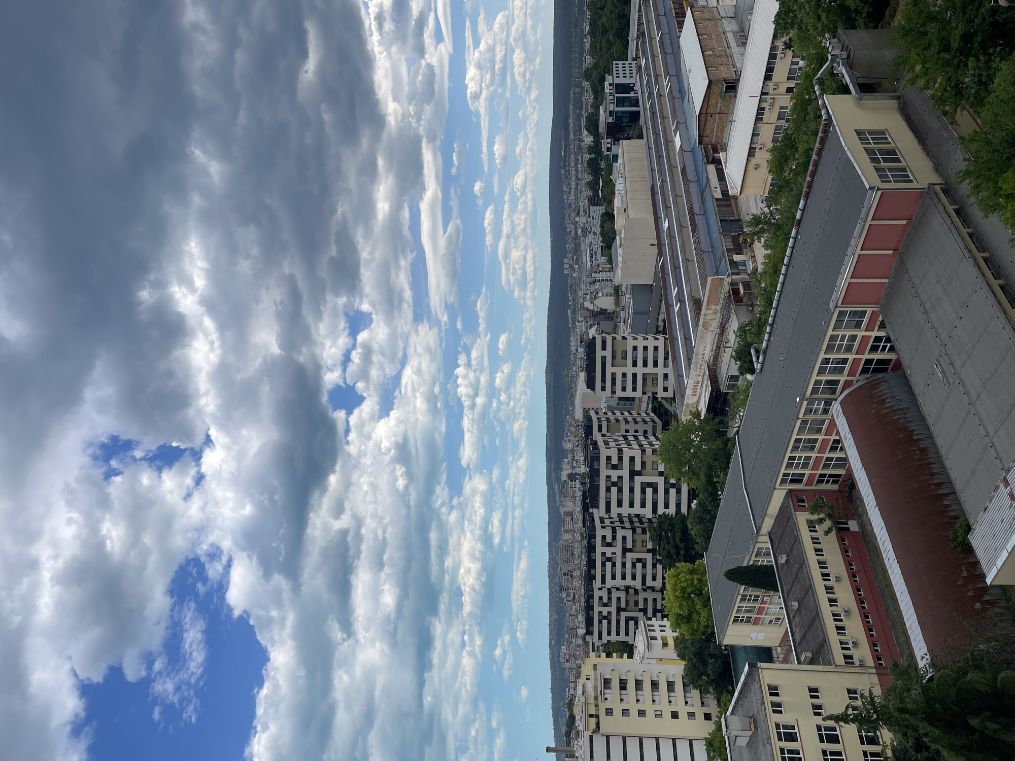 This is a high-angle, vertically-oriented shot of Varna, Bulgaria, featuring a cloudy sky covering much of the frame. The cityscape is visible on the right side, with various buildings of different heights and architectural styles, including some with modern patterns. There are signs of vegetation interspersed among the buildings. The sky is a mix of blues and grays due to the cloud cover. The orientation suggests that the photo was either taken from a tall building or rotated after capture.