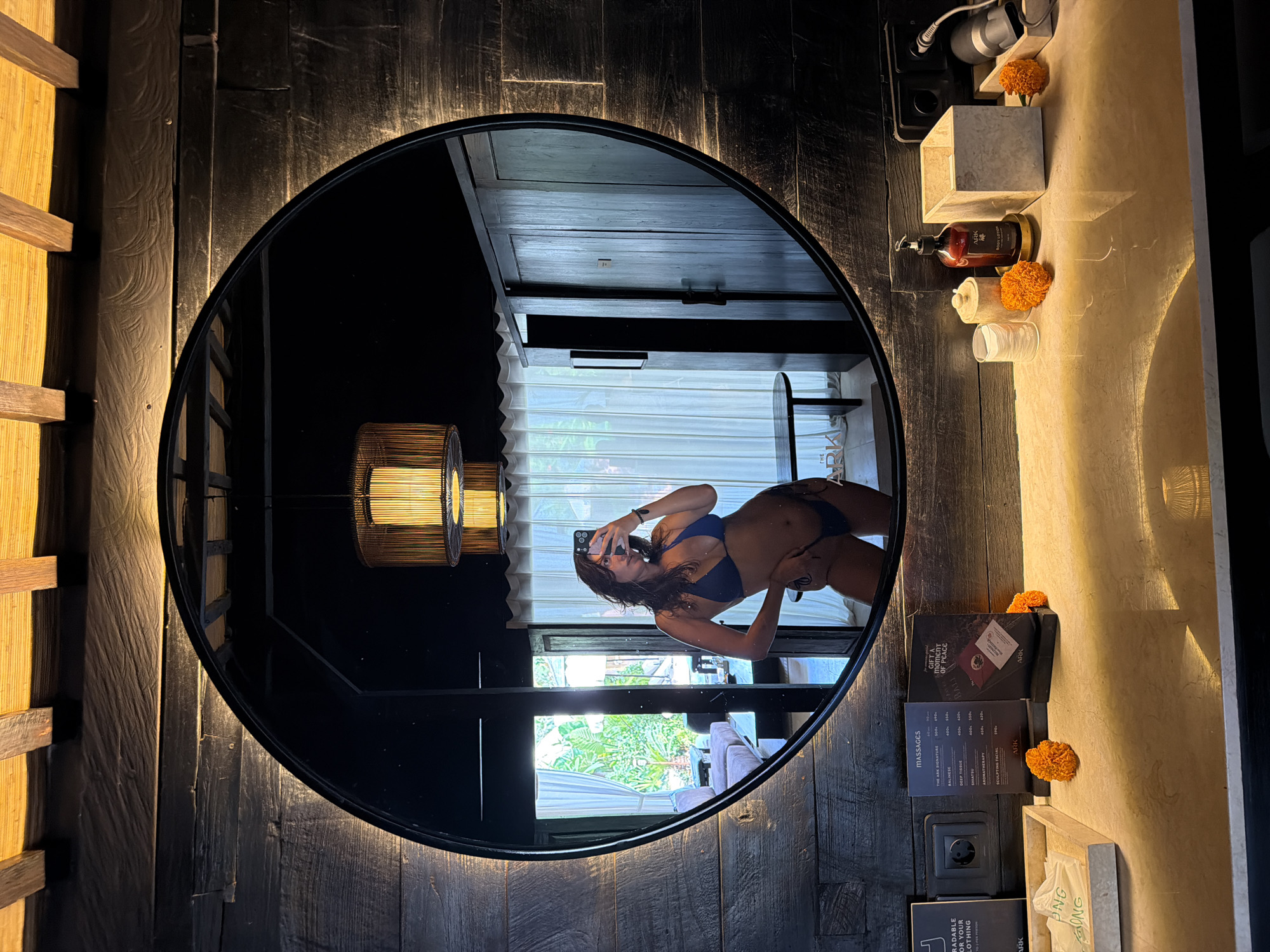 A woman in a blue bikini takes a mirror selfie in a dimly lit room with wooden walls and ceiling beams. She is standing in front of a large circular mirror, which reflects the room behind her. The reflection shows a large window with sheer curtains, a couch, a table, and a wooden wardrobe. On the counter in front of the mirror are several items, including a menu, toiletries, and decorative flowers. The scene appears to be in a hotel room or spa in Mengwi, Indonesia, given the "ARK" branding on some of the items and the overall aesthetic.