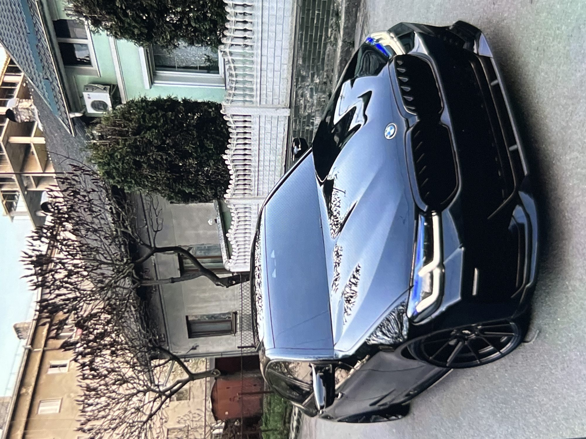 An angled, eye-level, front-view shot captures a black BMW sedan parked on a residential street. The car's sleek design is highlighted by its glossy finish and the blue accents in its headlights. Behind the car, a light green house with a dark green roof stands adorned with a white picket fence and manicured evergreen shrubs. Further in the background, a multi-story building under construction, with its exposed concrete framework, contrasts with the established residential structures. Bare trees with twiggy branches frame the scene, adding a touch of nature to the urban setting. The street is paved with asphalt, and the sky above is overcast, suggesting a cool or cloudy day. No people are visible in the frame. The overall impression is one of a quiet, suburban street.