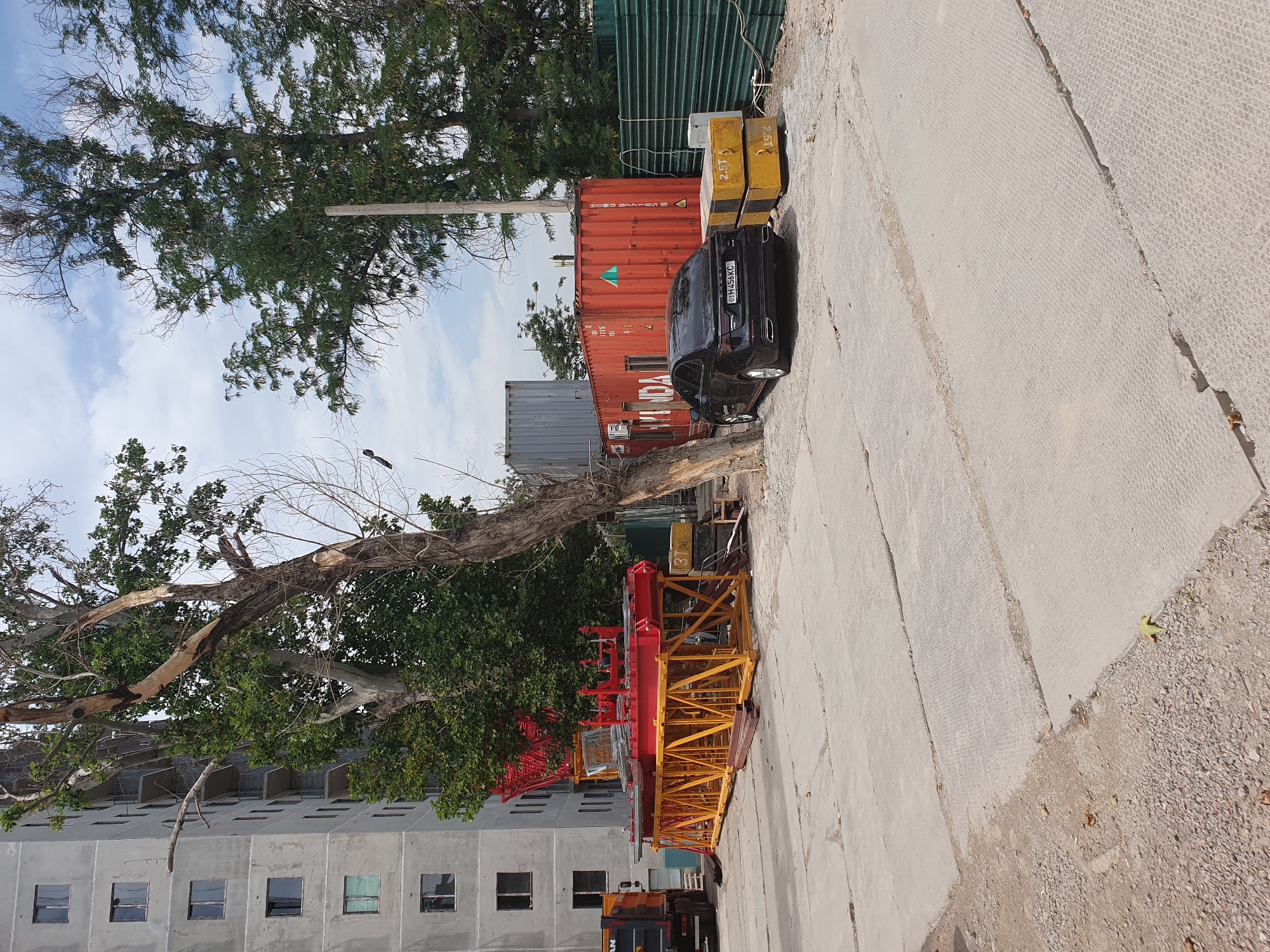 The image depicts an outdoor construction site in Tashkent, Uzbekistan, with a partially constructed multi-story building on the left. A large yellow and red crane structure is disassembled in the foreground. A black car is parked next to a stack of yellow concrete blocks, one marked "2.5T". Several shipping containers, one red and one grey, are visible in the background. Trees with dense foliage and some bare branches are interspersed throughout the scene, and the ground is covered with concrete slabs and gravel. The sky is partly cloudy.