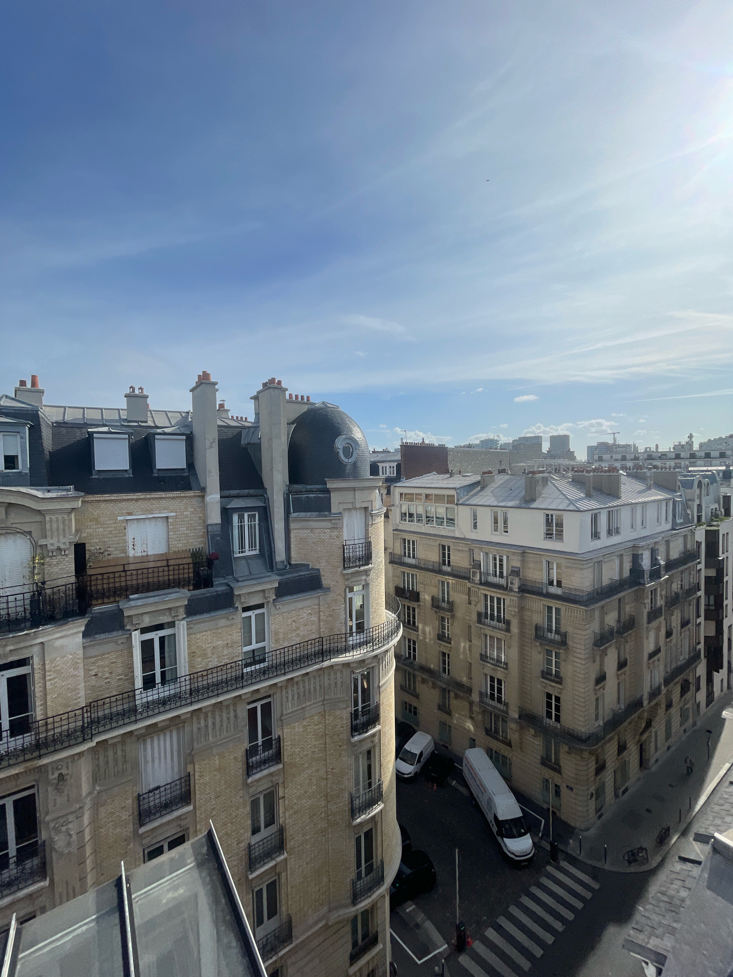 An aerial shot captures a sunny day in Paris, France. The image focuses on the intricate architecture of Parisian buildings, characterized by their mansard roofs, dormer windows, and ornate balconies. The prominent buildings in the foreground are made of light-colored brick, with dark metal railings adorning the balconies and windows. One building features a distinctive dark, domed structure on its roof.
Below, the street level reveals a crosswalk and several white vans. One van is parked diagonally across the street, its side displaying a logo with French text. Shadows from the buildings stretch across the pavement, indicating the sun's position. The sky is a clear blue with a few wispy clouds, and the sun glints brightly in the upper right corner. In the distance, more buildings of varying heights and architectural styles can be seen, forming the characteristic urban landscape of Paris. The scene appears to be a quiet street corner in a residential or mixed-use district.