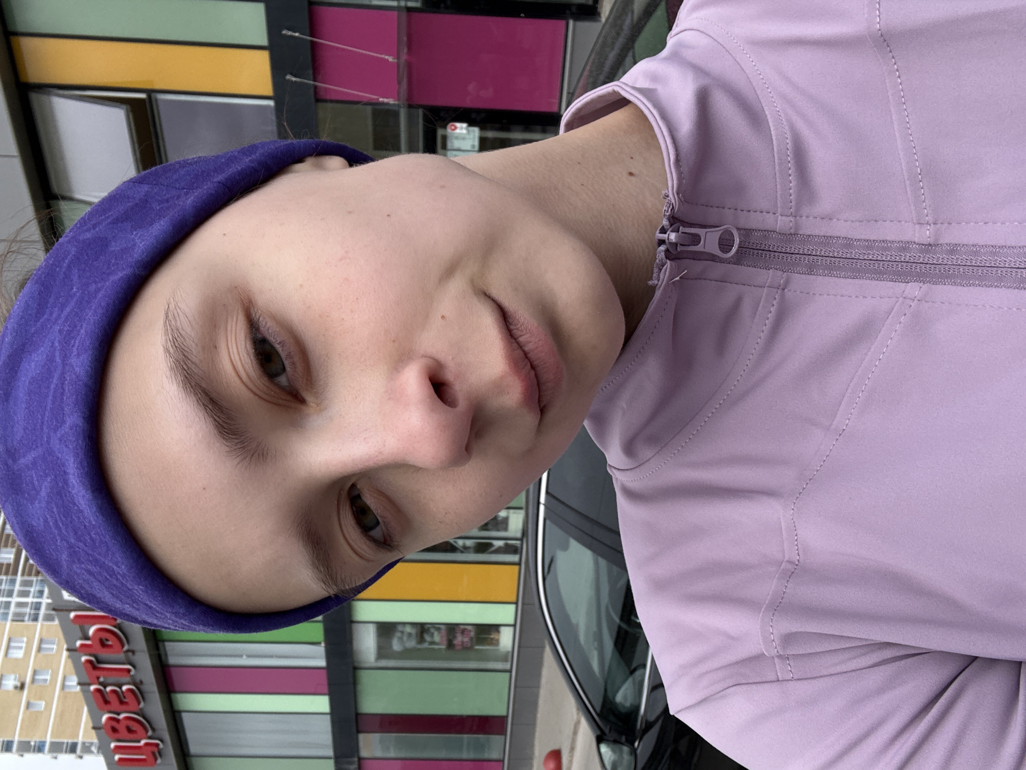 A young woman with fair skin and brown eyes, wearing a purple headband and a lavender-colored zippered jacket, is looking at the camera. The background includes a building with colorful panels (yellow, green, pink) and a sign with Russian letters reading "ЦВЕТЫ" (flowers). The side of a dark-colored car is partially visible on the right side of the frame. This photo appears to be taken in Perm, Russia, indicated by the Russian text on the storefront.