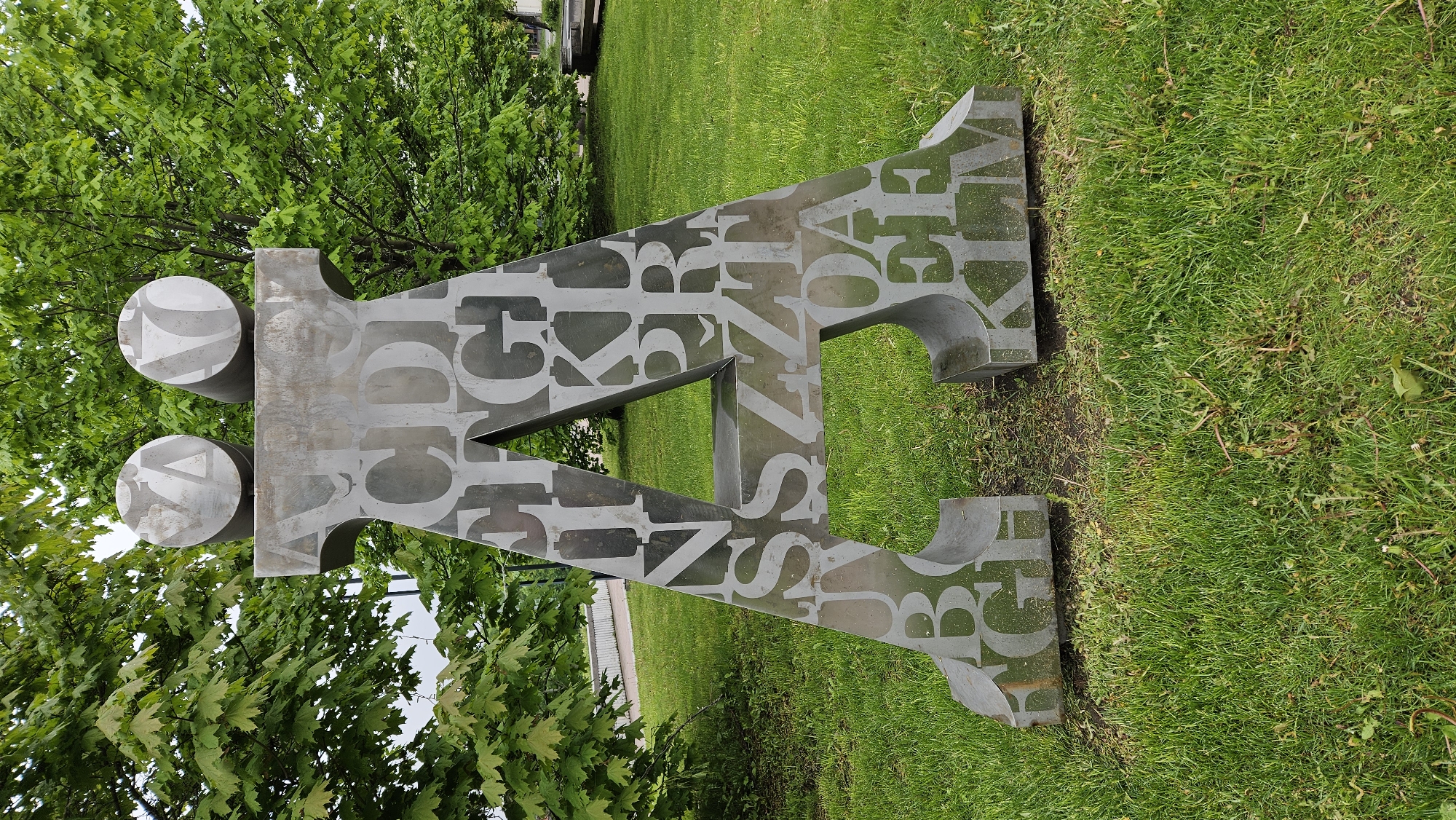 An outdoor sculpture featuring two circles above an angular shape is located in Unknown, Unknown, set against a backdrop of green trees and grass. The sculpture is a silver color, with lettering cut out of it.