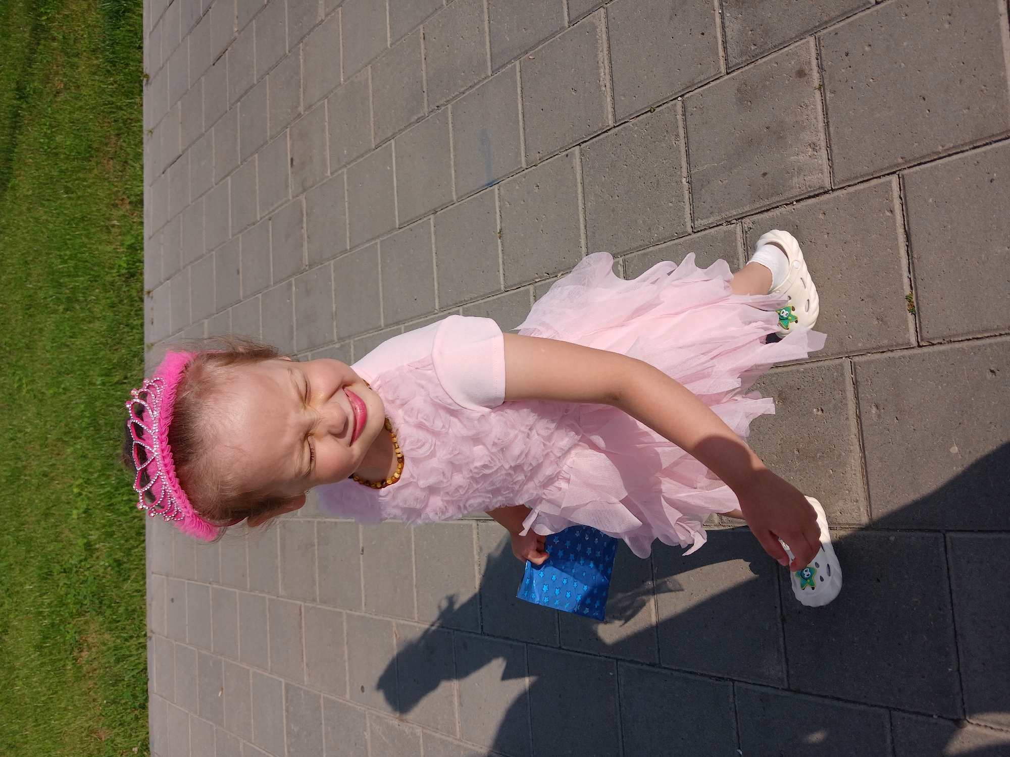 A young girl, dressed in a pink princess-like dress and a sparkly pink tiara, stands on a paved pathway next to a grassy area. She is holding a blue gift bag with white stars. Her eyes are closed, and she is sticking out her tongue, suggesting a playful expression. She wears white clogs with green charms. A shadow of a person is cast on the pathway. The location is Yekaterinburg, Russia.