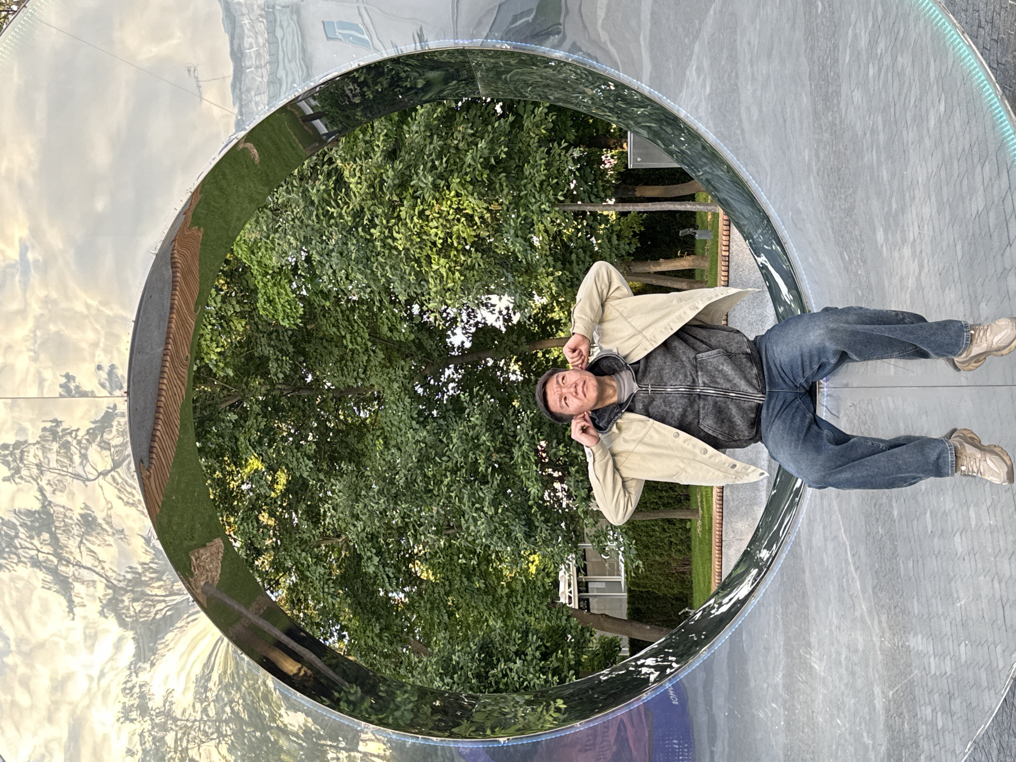 An East Asian man is depicted lying inside a large, circular, highly reflective metal art installation, likely located in Moscow, Russia. He is positioned on his back, with his head slightly raised and turned towards the camera, wearing a light beige jacket over a dark grey hooded top, blue jeans, and light-colored sneakers. His right hand is near his ear, and his expression is relaxed and contemplative.
The metallic structure forms a significant part of the composition, creating a reflective tunnel or archway. Its polished surface mirrors the surrounding environment, including a bright sky with clouds, distant buildings, and lush green trees that are also visible directly through the opening of the circle. The reflections create abstract and distorted patterns across the shiny surface. A subtle strip of blue-green lights is visible along the outer edge of the circle on the right side. The ground beneath the man, forming the base of the installation, appears to be a light grey, textured surface.
The scene takes place outdoors during what appears to be daytime, possibly late afternoon, given the soft, ambient light. The man seems to be casually posing or relaxing within the art piece, inviting interaction with the unique structure. While the general location is Moscow, Russia, specific landmarks are not distinctly visible, although reflections hint at urban surroundings. Faint, partially obscured text can be seen on one of the reflective surfaces, but it is not clearly legible.