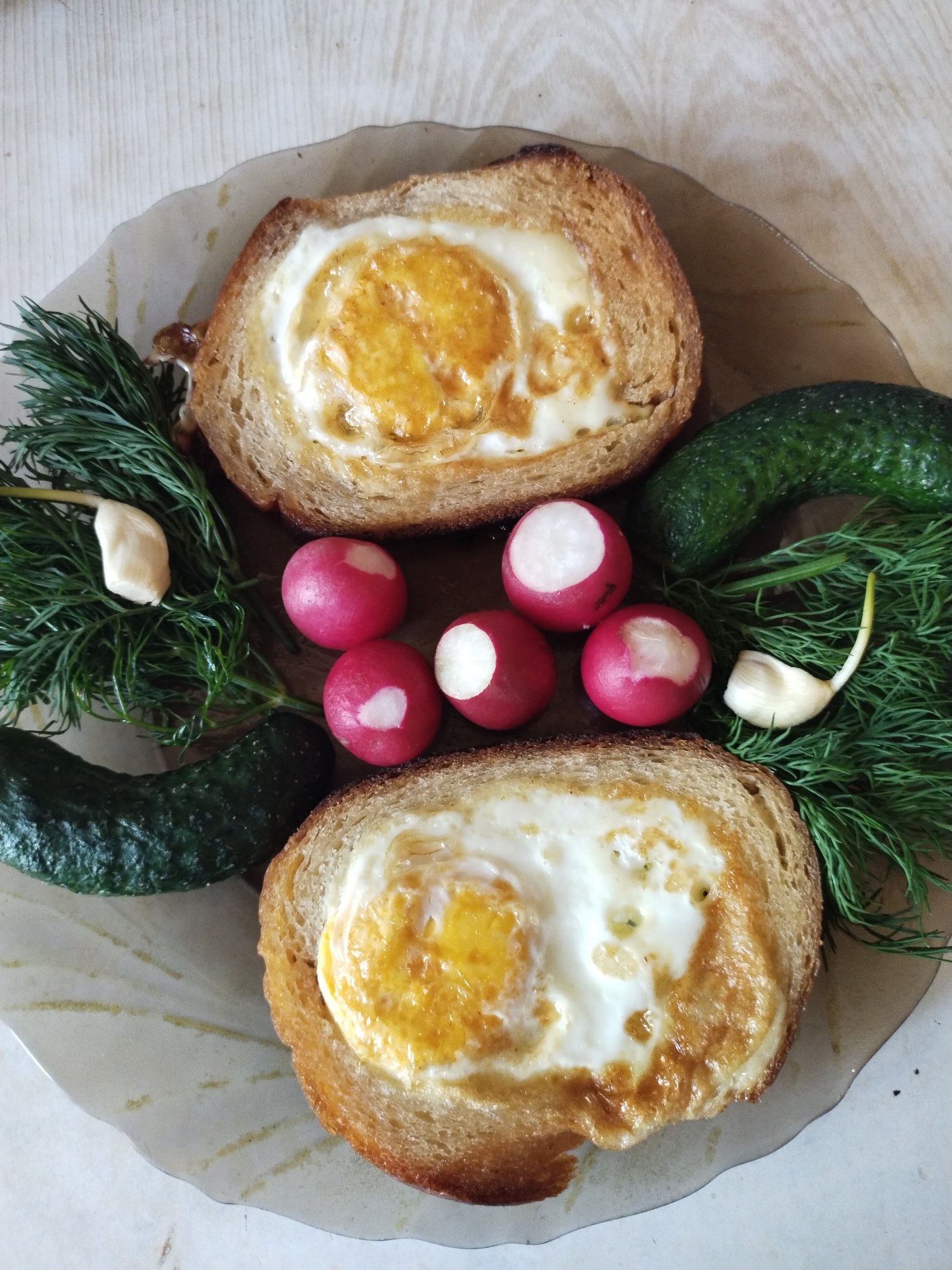 A close-up, overhead shot displays a plate of food on a wooden surface. The plate contains two slices of toast, each with a fried egg cooked inside a cutout. Both eggs appear to be over-easy, with slightly runny yolks and set whites. The toast is golden brown and appears slightly toasted. Surrounding the toast are several sprigs of fresh dill, two small cucumbers, and five radishes. Two cloves of garlic, peeled and white, are also present among the dill. The lighting is natural, casting soft shadows. The overall presentation suggests a simple, homemade meal. This image was taken in the city of Horishni Plavni, Ukraine.
