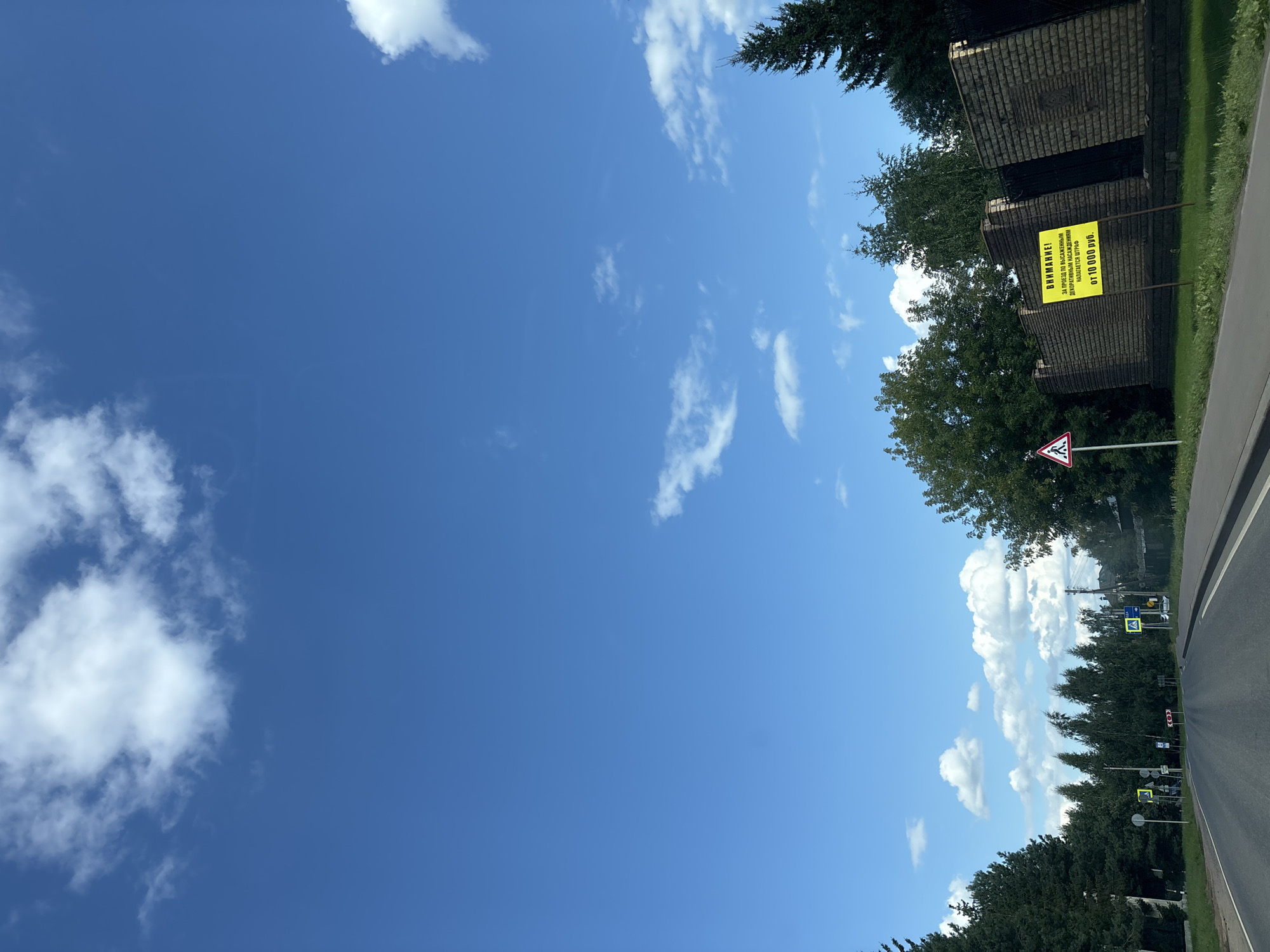 The image captures a road scene on a sunny day under a blue sky with scattered white clouds. On the right side of the road, there is a brick wall and two yellow signs. The left sign states "ВНИМАНИЕ!" (ATTENTION!) and the right sign reads "ЗА ПРОЕЗД ПО ВЫСАЖЕННЫМ ДЕКОРАТИВНЫМ НАСАЖДЕНИЯМ НАЛАГАЕТСЯ ШТРАФ ОТ 10 000 РУБ." (For driving on planted decorative landscaping, a fine of from 10,000 rubles is imposed.). The road curves to the left, bordered by a curb. On the left side of the road, there are trees and several traffic signs, including a pedestrian crossing sign. In the distance, more trees and some buildings are visible. The overall environment suggests a road within a populated area, possibly a village or suburb, given the presence of decorative landscaping and residential structures. The image does not depict any people or specific actions, focusing on the environmental and infrastructural elements. The mention of "Millennium Park, Russia" as a location is not directly ascertainable from the visual information provided in the image.