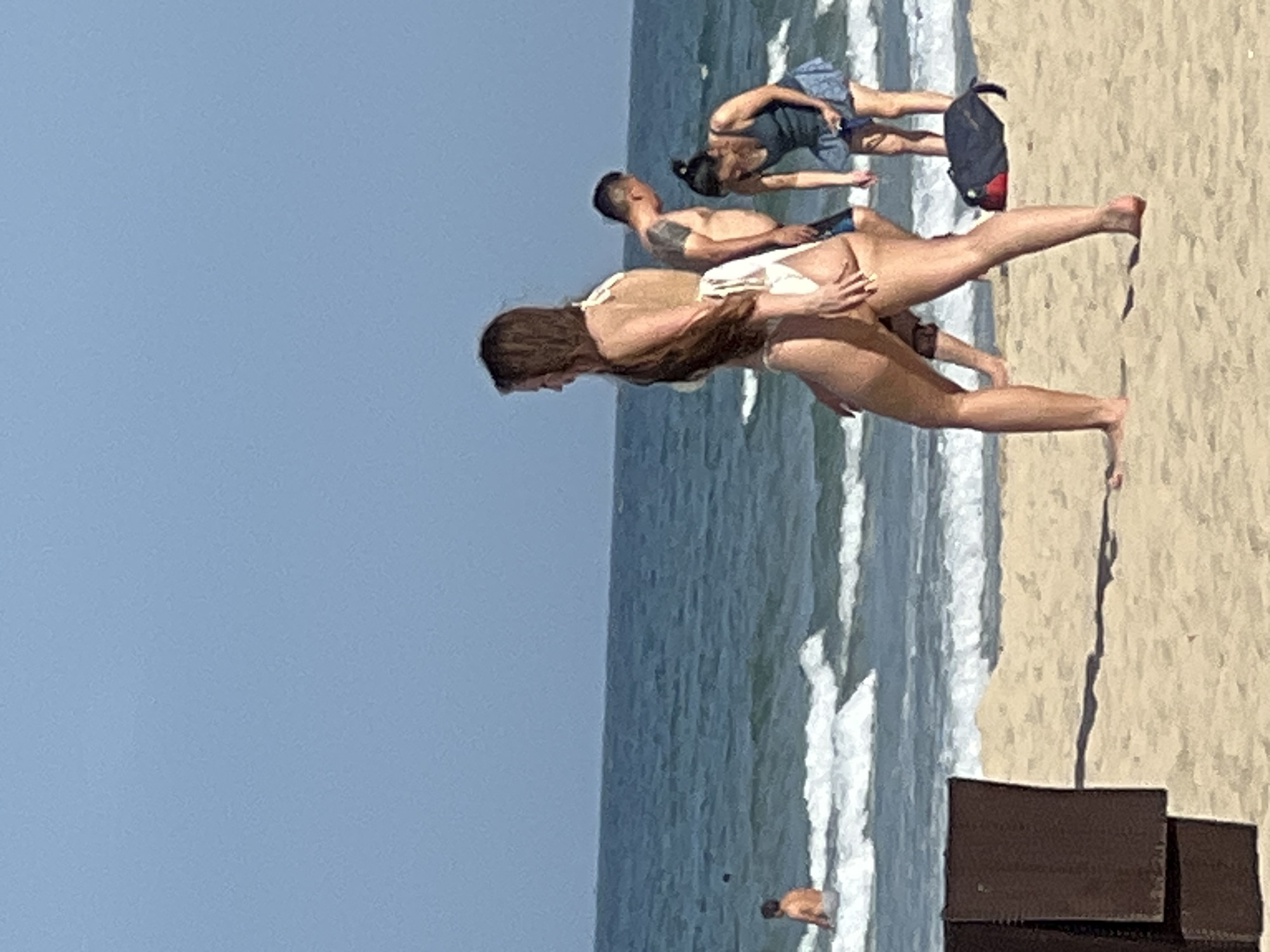 The image depicts a scene on a sandy beach in Đà Nẵng, Vietnam. The ocean, with moderate waves and white foam, occupies the background, under a clear, light blue sky. In the foreground, the sandy beach is visible, with a dark brown object, possibly a beach chair or lounge, positioned on the left side.
Several individuals are present. In the mid-ground, a woman with long brown hair is walking towards the right, wearing a light-colored bikini. Behind her and to the right, a man with short dark hair and a tattoo on his shoulder, wearing dark swimming trunks, is also present. Further to the right, a woman with dark hair pulled back in a bun is bending over, wearing a dark blue tank top and a blue skirt. A dark bag with red accents lies on the sand near her. In the distance, on the left side of the frame, a person is standing in the ocean, partially obscured by waves. The overall atmosphere suggests a casual day at the beach.