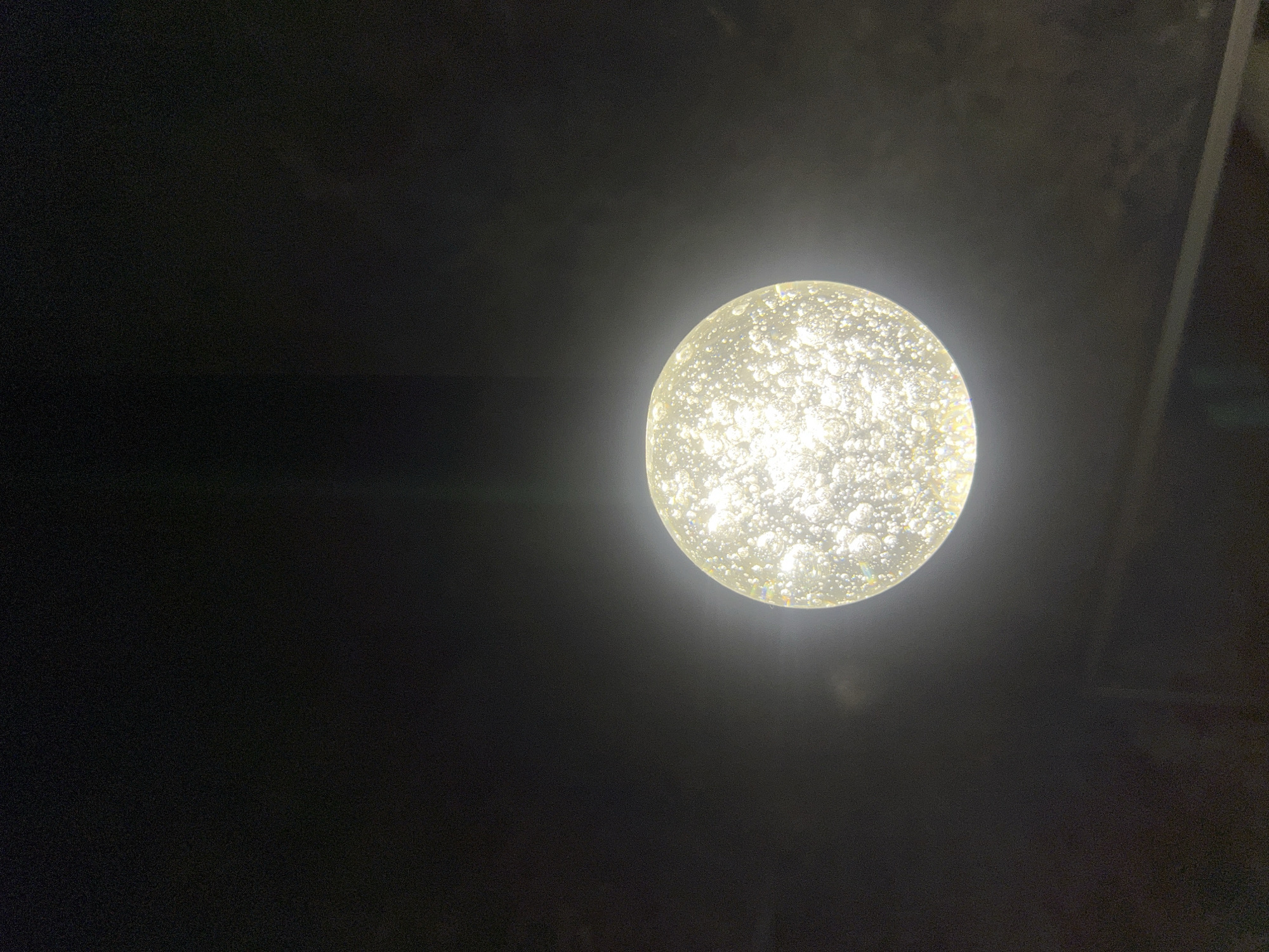 A single, illuminated, orb-shaped light fixture is the central subject of this image, set against a dark, ambiguous background. The orb is textured with numerous small, irregular bubbles or inclusions, which catch and refract the light, giving it a sparkling, ethereal quality. The light emanating from the fixture is bright and appears to be a warm, white tone, casting a soft glow that illuminates the immediate surroundings. The background is predominantly black, with subtle variations in shade suggesting depth or texture, possibly a ceiling or a wall. There are faint linear elements visible in the upper right portion of the frame, which might indicate structural features of the setting. The overall impression is one of a dimly lit environment, possibly indoors at night, where the light fixture is the primary source of illumination. There is no indication of people or specific activities taking place. The visual focus is on the abstract beauty and texture of the illuminated orb. No text is visible in the image.