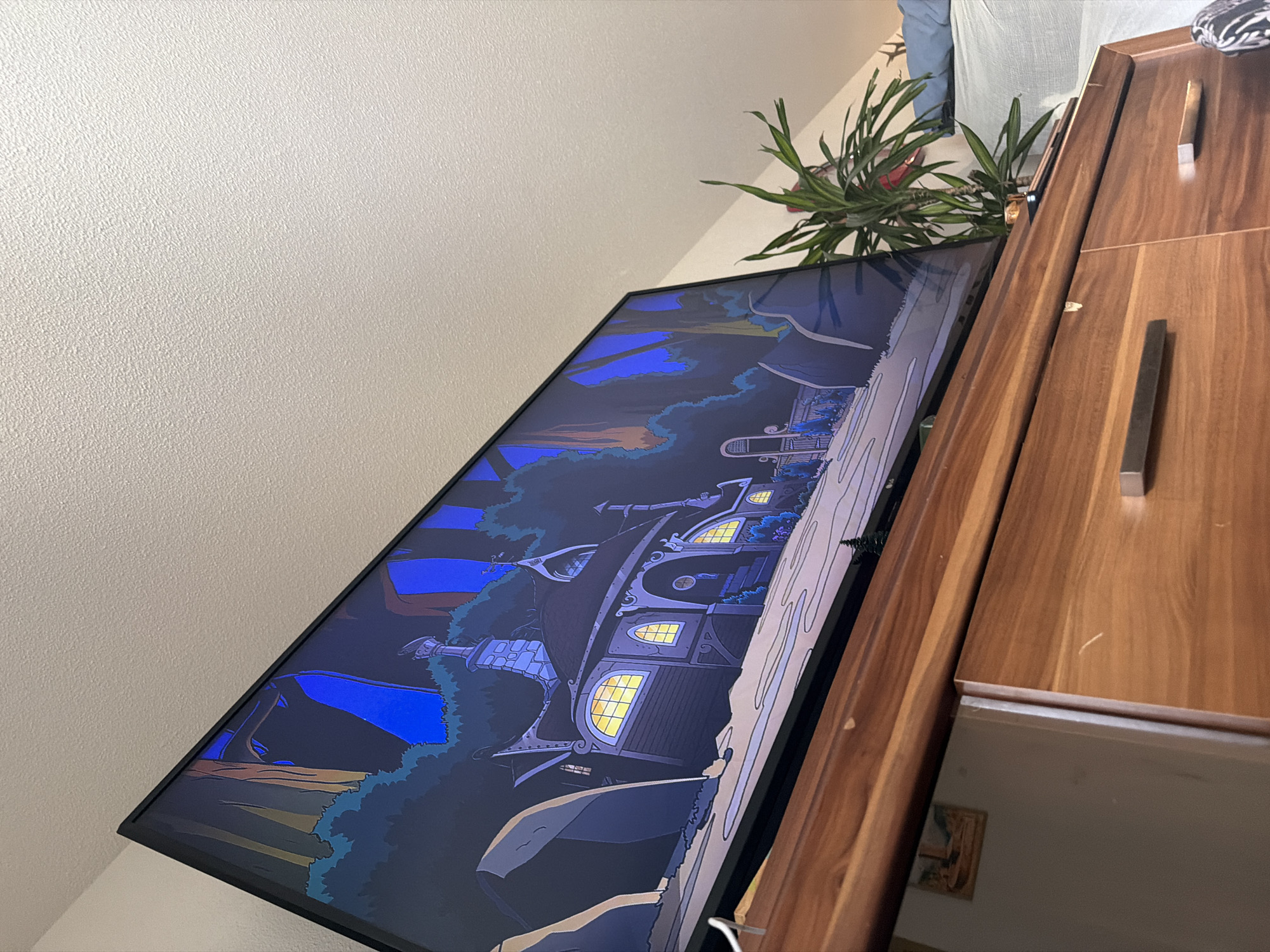 The image, taken indoors in what appears to be a living space or bedroom in Strasbourg, France, is rotated 90 degrees counter-clockwise. A large, black-framed flat-screen monitor or television dominates the central and left portion of the view, tilted slightly upwards. The monitor displays an animated scene or artwork, depicting a whimsical, multi-story house or castle with numerous glowing yellow windows, suggesting a fantastical setting at twilight or night. The sky in the artwork is a deep blue, accented with lighter blue and subtle orange hues, while dark green and blue foliage surrounds the structure. Several blue banners or flags are also visible in the foreground of the digital image.
Adjacent to and partially obscuring the monitor on the right is a robust, reddish-brown wooden cabinet or wardrobe, exhibiting a distinct wood grain and equipped with modern, rectangular metallic handles. Above and behind the monitor, a healthy potted plant with long, spiky green leaves adds greenery to the indoor environment. The background on the left consists of a uniformly light-colored wall with a textured finish. In the lower right, a mirrored surface reflects a small portion of the room, including a faint image of what appears to be a framed item. No individuals are present in the scene, and no discernible text is visible anywhere. The overall lighting is soft, emphasizing the textures of the room's elements.