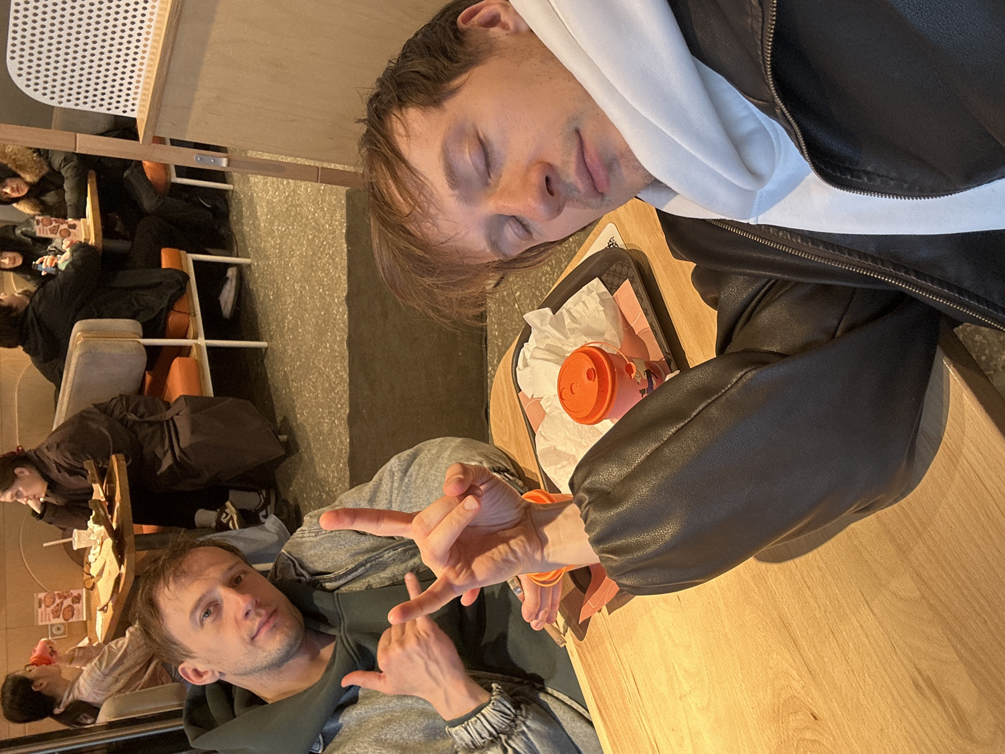 The image depicts an indoor scene in Murino, Russia, likely a cafe or fast-food restaurant. Two men are prominently featured at separate wooden tables, with other patrons visible in the background.
On the right side of the frame, a man with brown hair, wearing a black leather jacket over a white hoodie, is resting his head on a wooden table with his eyes closed, appearing to be asleep or deeply relaxed. Beside him on the table is a black serving tray holding an orange plastic cup and crumpled white paper napkins.
To the left, another man with short brown hair and light stubble, dressed in a grey zippered hoodie over a darker shirt, looks directly towards the camera. He is making a "shaka" or "hang loose" hand gesture with his right hand, extending his thumb and pinky finger while curling the others. A similar, less defined gesture is made with his left hand.
In the background, several other individuals are seated at similar light-colored wooden tables, suggesting a busy public dining area. Some are looking at their phones, while others appear to be conversing. One woman is visible in a dark coat with a fur-lined hood. The floor is a speckled, light-colored material, possibly concrete or tile. No discernible text is clearly visible in the image. The time of day is not evident from the indoor lighting.