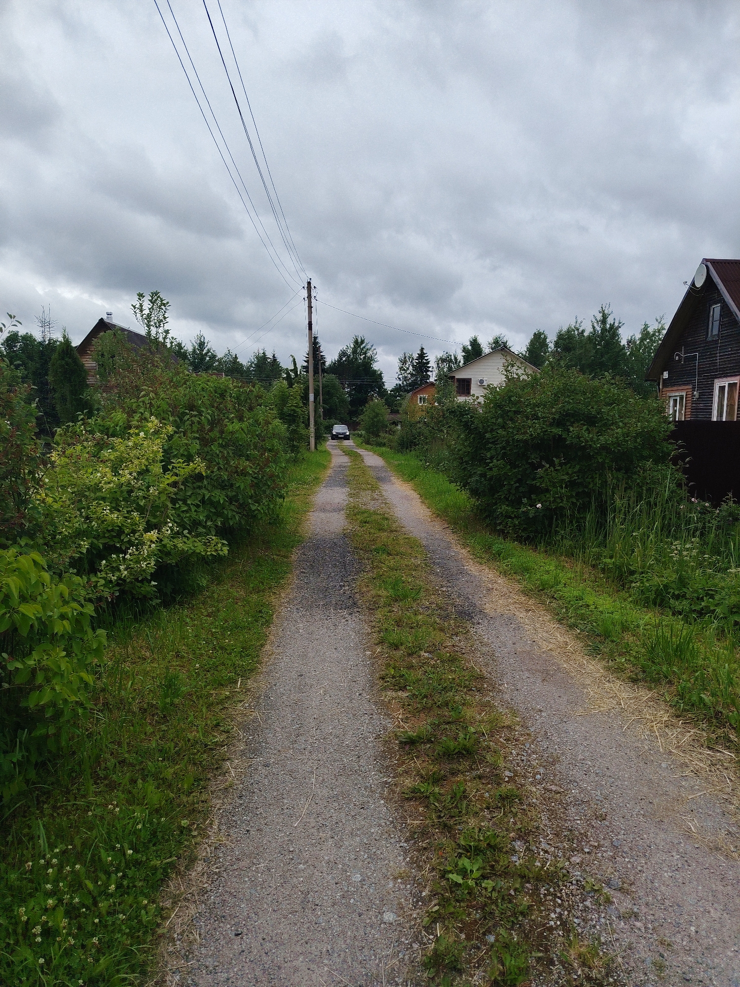 An outdoor, eye-level, medium shot depicts a gravel road leading into a village, under a cloudy sky. On the right side of the road, a dark wooden house with a dark red roof and a satellite dish is visible. Further down the road, a dark-colored SUV is parked. The left side of the road is bordered by lush green foliage, including trees and bushes, and a few small white flowers. The road itself is a narrow, unpaved path, suggesting a rural or suburban setting. The scene is devoid of any human presence or activity, creating a sense of quietude. The overall atmosphere is overcast, with soft, diffused light.