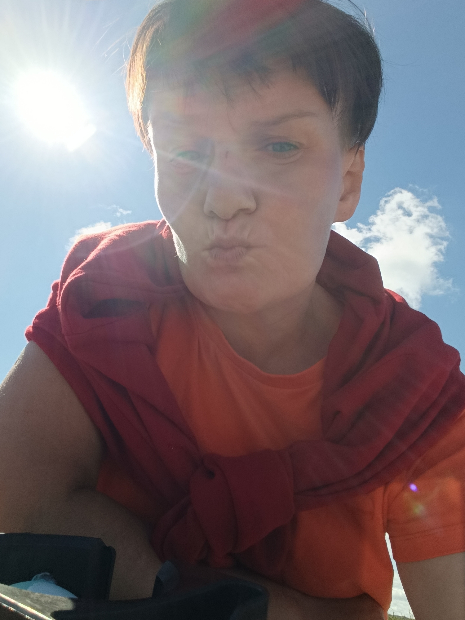 A woman with short, dark hair and blue eyes is captured in a low-angle, outdoor selfie. She is wearing an orange t-shirt with a red sweater casually draped around her shoulders. Her mouth is puckered in a kiss. The bright sun is visible in the upper left corner of the frame, creating lens flare and casting a glare on her face. The background consists of a bright blue sky with scattered white clouds. The overall impression is a candid, sunny day moment. The image does not contain any discernible elements that would allow for identification of a specific location in the city of Фоки, Russia.
