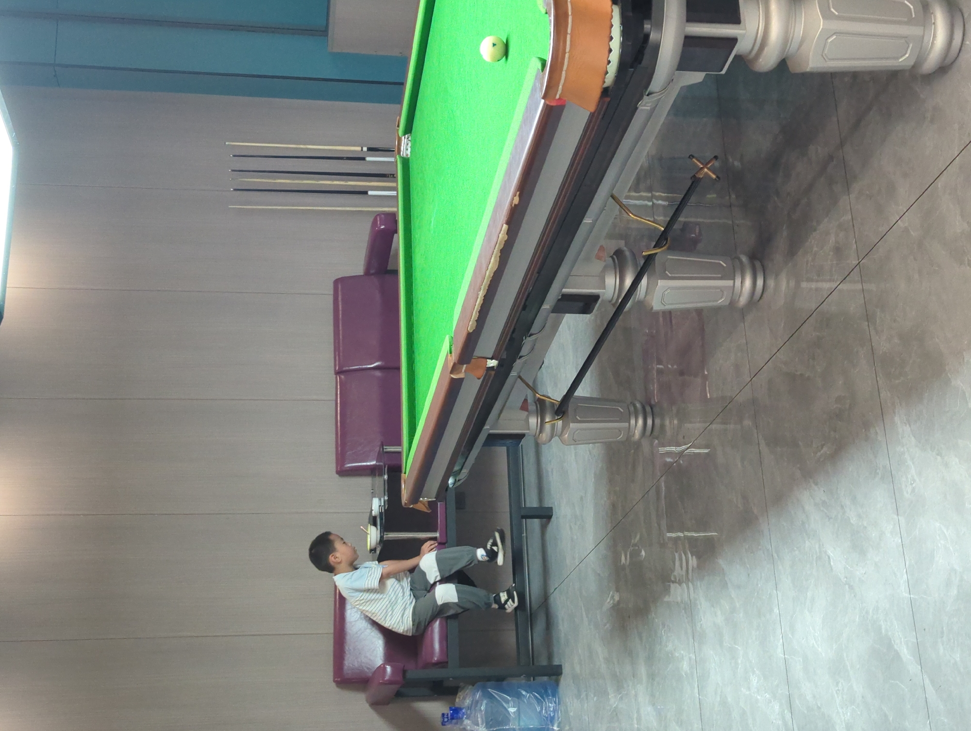 The image depicts an indoor setting that appears to be a billiard hall or a similar recreational space. In the foreground, a portion of a green billiard table is visible, featuring its textured felt surface, wooden rails with some wear, and ornate silver legs. A single white billiard ball rests on the table. To the left of the table, a young boy is seated on a dark purple cushioned bench. He is wearing a striped t-shirt and grey pants, and is looking down at a small bowl placed on a dark table in front of him. Behind the bench, more of the purple seating continues, forming a sectional sofa. A tall dispenser of bottled water is positioned to the left of the bench. In the background, several billiard cues are vertically arranged. The walls are paneled with a light brown, wood-grain material. The floor is tiled with a polished, grey, marble-like pattern, reflecting the overhead lighting. The overall impression is that of a dimly lit, quiet establishment.