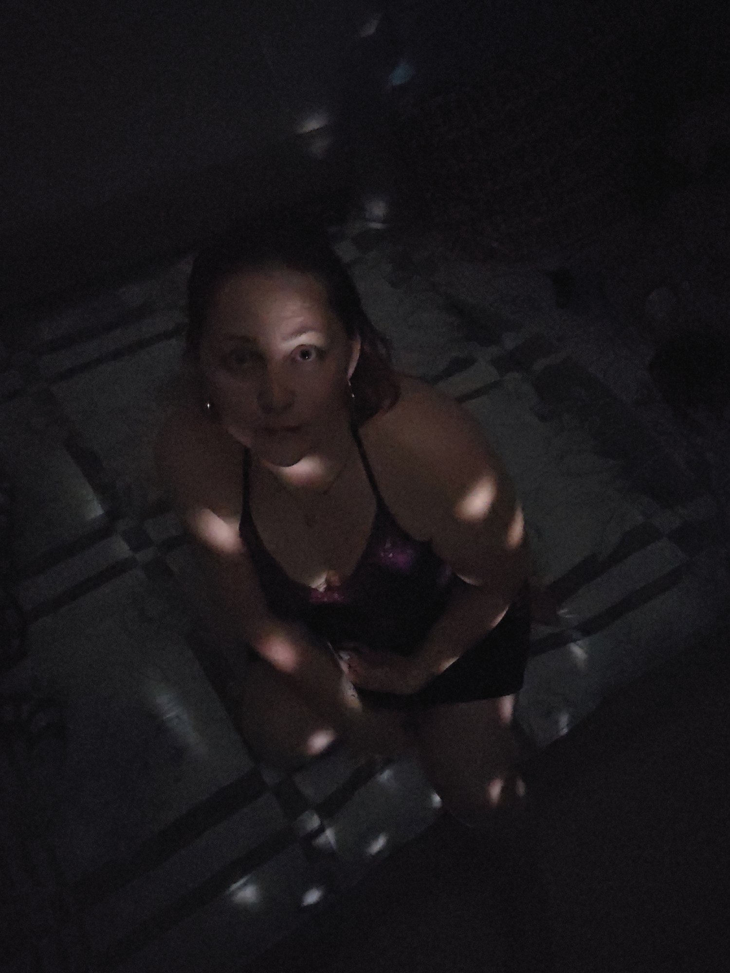 The image shows a person, likely female, kneeling or crouching in a dimly lit indoor space. The subject has light skin and reddish or brownish hair, and is looking directly upwards towards the camera with a neutral expression. She is wearing a dark-colored top, possibly a swimsuit or camisole, with some visible pattern on the bust area, and dark shorts. Small earrings are visible.
The scene is characterized by dramatic, high-contrast lighting. Bright, circular or elliptical patches of light are cast onto the subject's face, shoulders, arms, and legs, creating a dappled effect. Similar light patterns are visible on the dark floor, which appears to have a subtle grid-like or striped pattern. The rest of the background and surroundings are plunged into deep shadow, suggesting very low ambient light, possibly at night or in a dedicated indoor setting. The person appears to be posing for the photograph. No text is visible in the image.