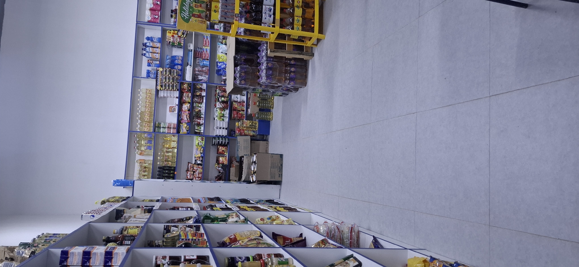 The image shows the interior of a small grocery store or market located in Arys, Kazakhstan. The store features white shelving units filled with various food products and household items. On the left, shelves are stocked with packaged goods, bottles, and jars. On the right, larger shelves display bottles of cooking oil, cleaning supplies, and other packaged items. A yellow display stand is visible in the foreground, holding bottles of drinks. The floor is tiled in a light grey color. The overall environment is organized and well-lit, typical of a retail setting. There are no people visible in the image.