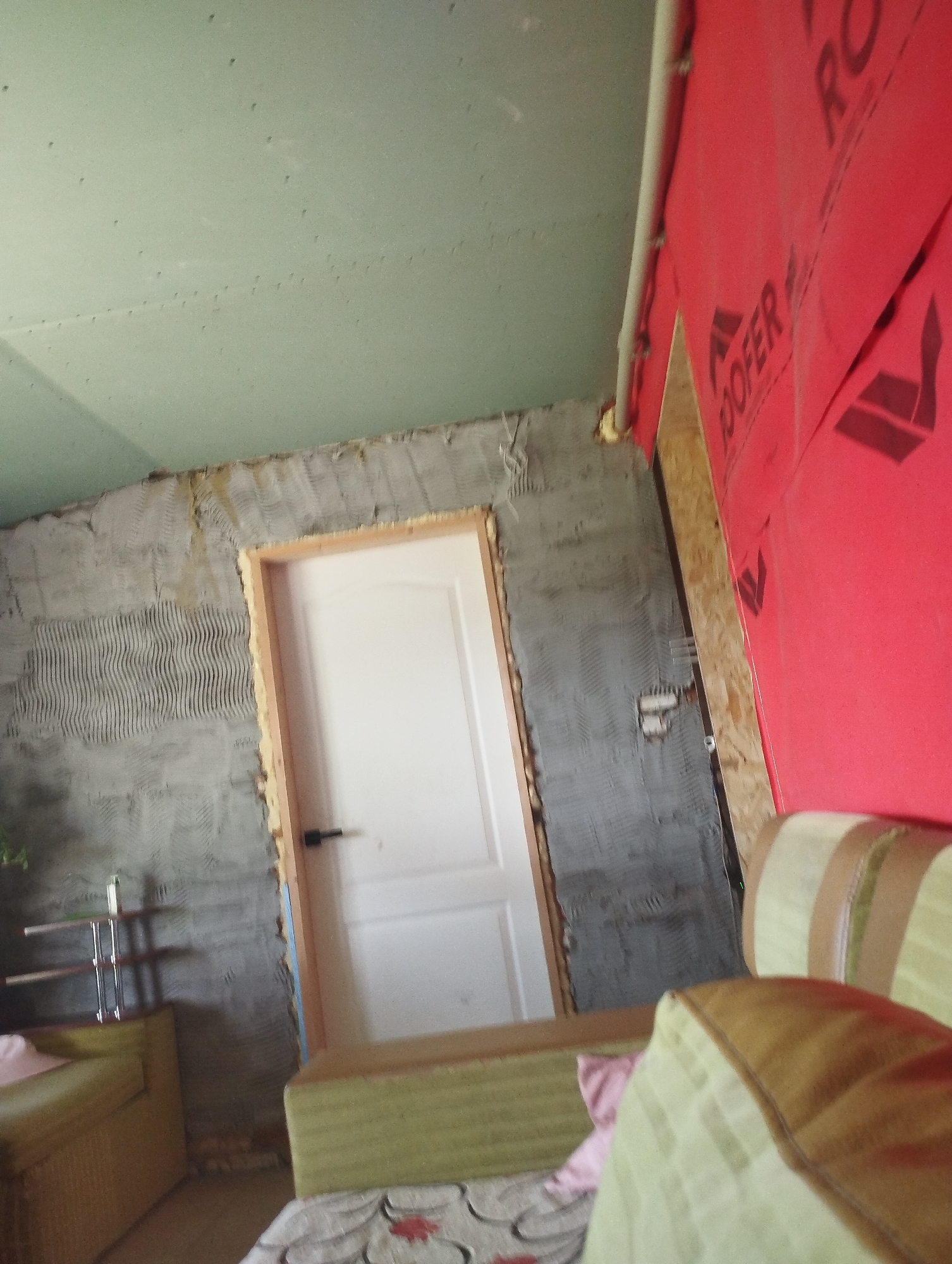 The image depicts an indoor scene, possibly a room under renovation, in Ustyvytsia, Ukraine. A partially plastered wall with a visible doorframe and a white door with a black handle is centrally located. To the right of the door, a wall is covered with red construction material featuring black lettering that appears to spell "ROOFER". To the left of the door, another wall has a textured gray plaster finish. In the foreground, a light green sofa with brown and light green striped cushions is visible, with another section of a sofa or chair in front of it. A metal shelf unit with at least two shelves is also present to the left, with what appears to be a bottle on the top shelf. The ceiling is a light green color with visible imperfections or holes. A light gray pipe runs vertically along the wall to the right of the door. The scene suggests ongoing construction or repair work.