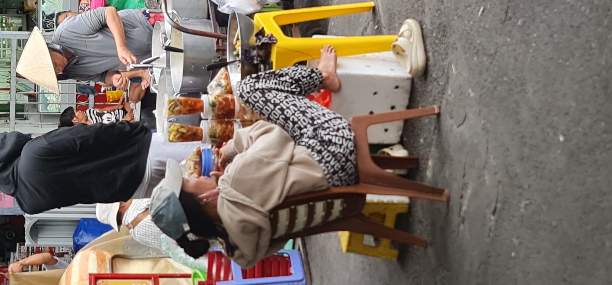 The image depicts a street scene in Đà Nẵng, Vietnam. A person is reclining on a brown plastic chair and resting on a white block, with their bare feet propped up on a yellow chair. The person is wearing patterned pants and a light-colored hooded sweatshirt. Next to them are several containers of food or beverages. Another person wearing a conical Asian hat is standing near metal pots and appears to be preparing food. A woman is lying down behind the man. A third person wearing a white hat is located at the bottom of the image. The background includes storefronts and a gray concrete street.