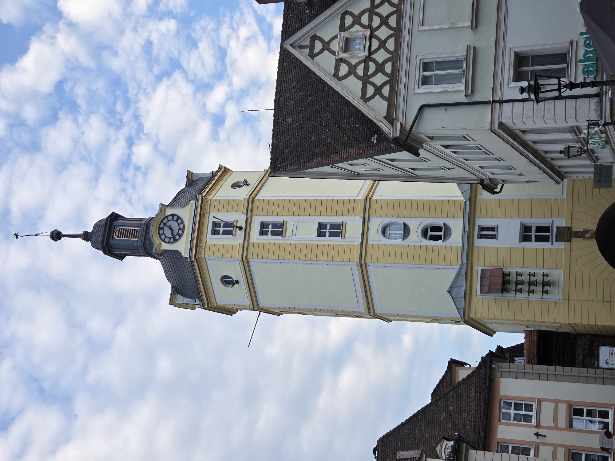 A tall, pale yellow tower with a dark gray, conical roof and a weather vane stands against a partly cloudy sky. The tower has a clock on one side and several rows of bells mounted on its facade. To the right of the tower is a light blue building with a dark gray, tiled roof and decorative patterns on its gables. To the left of the tower is a building with a pale pink and white facade, also with a dark gray, tiled roof. The scene appears to be an urban setting in Ansbach, Germany, likely a town square or street, given the presence of multiple buildings and architectural styles.