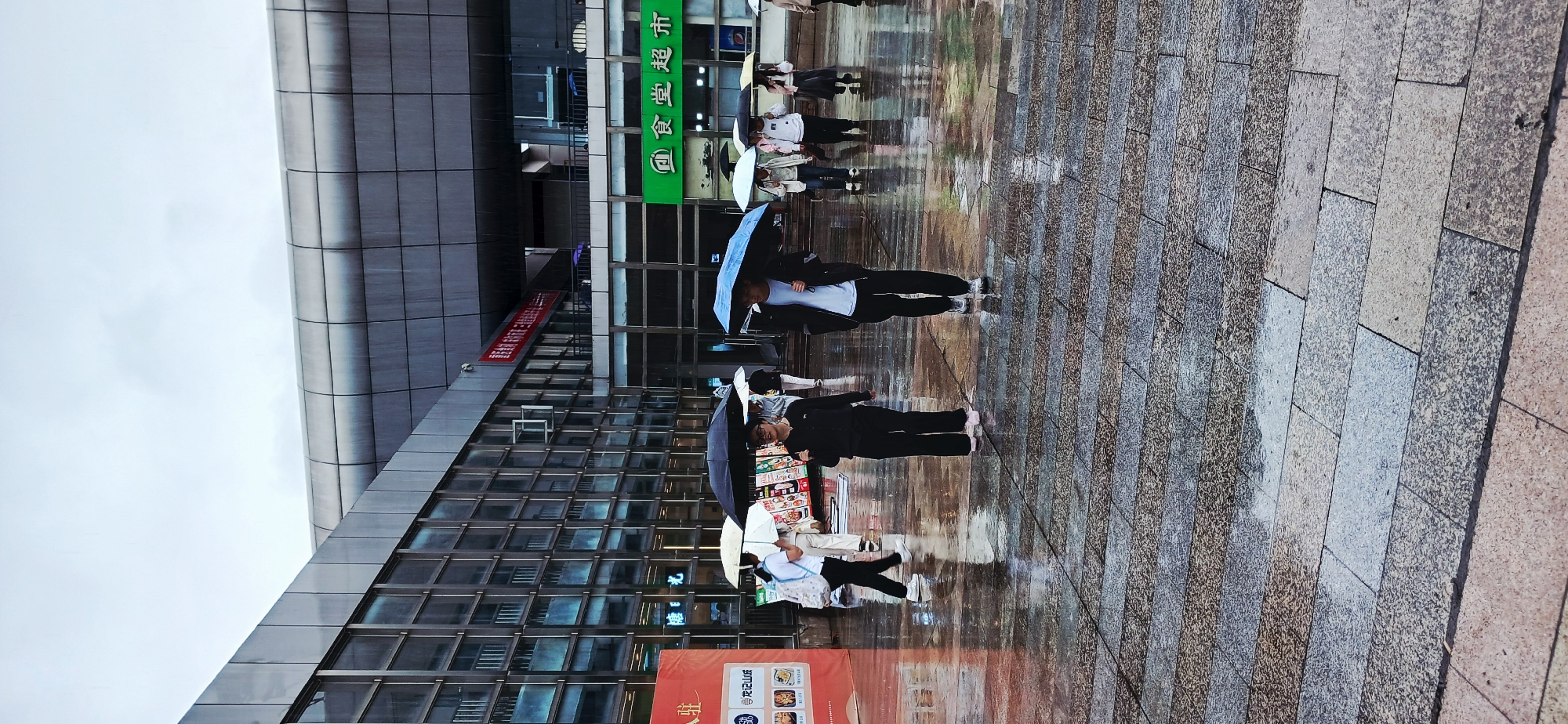 The image captures an outdoor urban scene in what appears to be a commercial or public area, likely in Chongqing High-tech Zone, China, given the visible text and provided context. The weather is overcast, and the ground is wet and reflective from recent rain, with several people holding umbrellas.
The foreground features a paved area with rectangular, textured tiles, showing visible puddles and reflections. Several individuals are walking across this area, generally moving from left to right. They are dressed in casual attire. At least three people are clearly holding umbrellas: one with a blue umbrella, one with a white umbrella, and another with a black umbrella. Their reflections are visible on the wet ground.
In the background, there are modern buildings with multi-story facades. One building on the left has a sleek, metallic, possibly curved structure. The main building in the center features large glass windows and horizontal paneling. A prominent green sign with Chinese characters "食堂超市" (Shítáng Chāoshì), meaning "Canteen Supermarket," is visible on this building. Below it, on the lower level, another red sign has partially visible Chinese characters, including "重庆" (Chóngqìng), confirming the location context. Other smaller signs and banners with indistinguishable text are also present. The sky is a uniform, bright grey, indicative of an overcast day.
